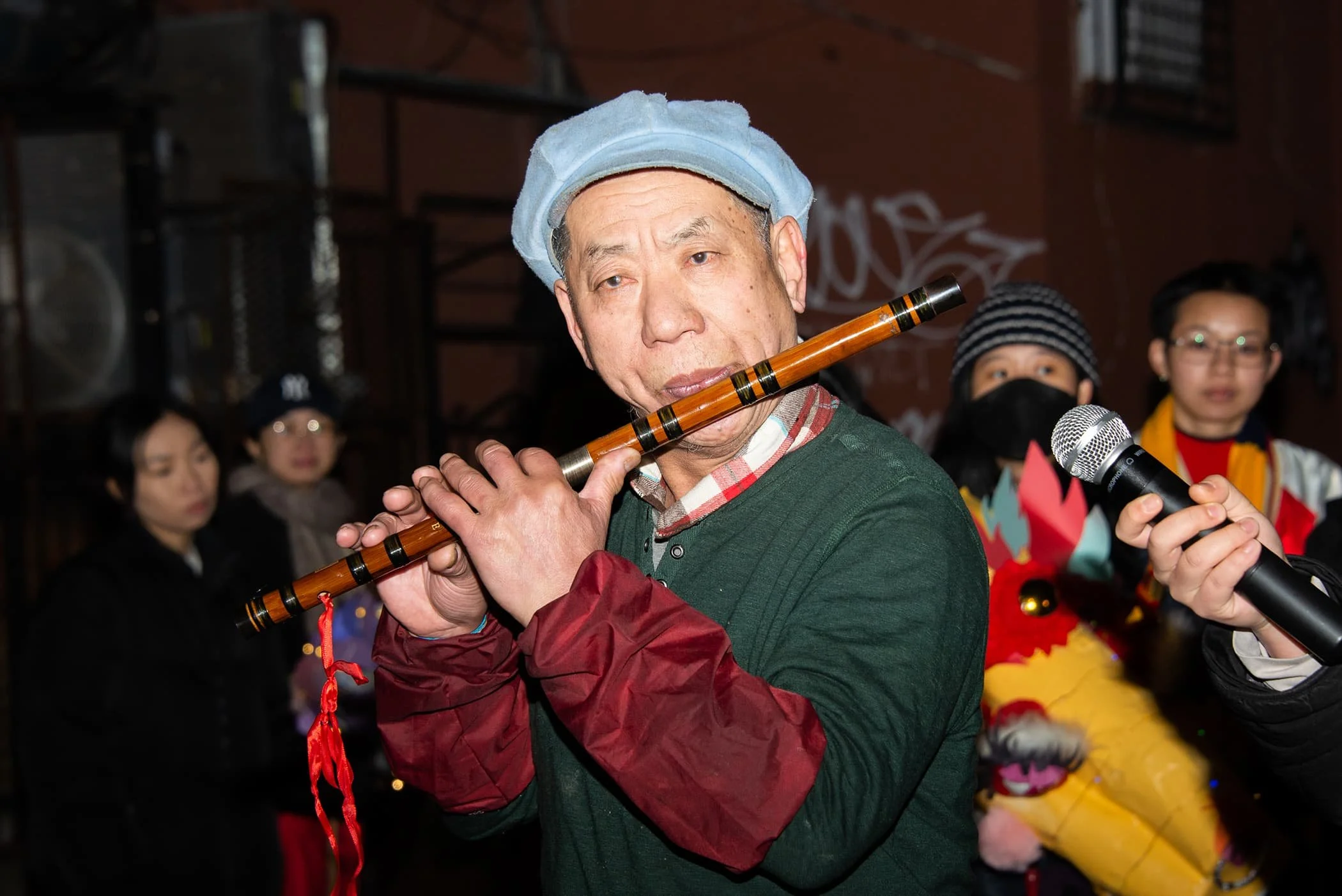 An elderly man in a blue hair net is playing a traditional bamboo flute, surrounded by a group of people, some wearing masks, at a cultural event or celebration.