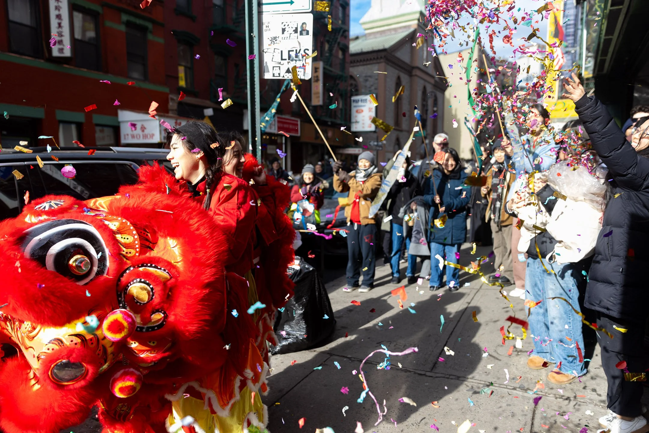 People celebrating with a lion dance, confetti, and festive clothing during a parade in a city street.