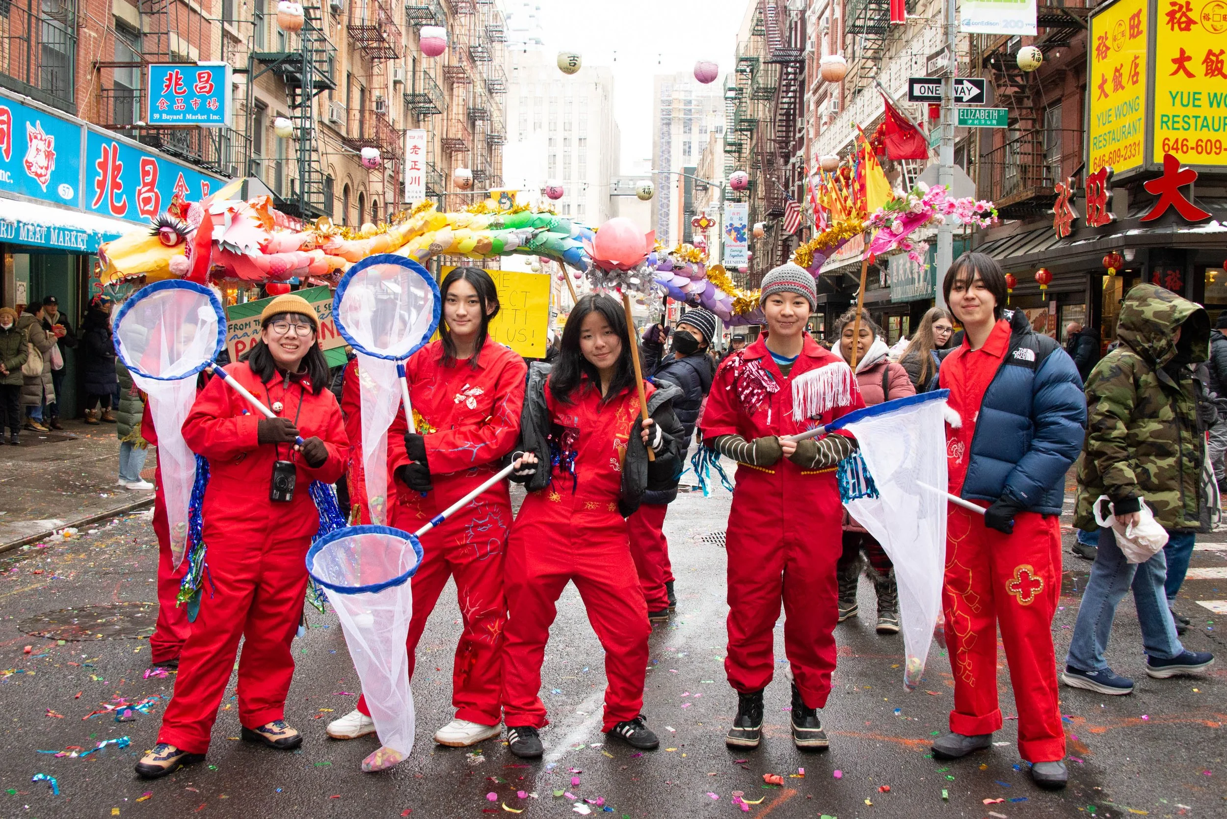 Group of women in red costumes holding butterfly nets standing in a street during a festival, with colorful lanterns and decorations overhead, and a crowd of people around.