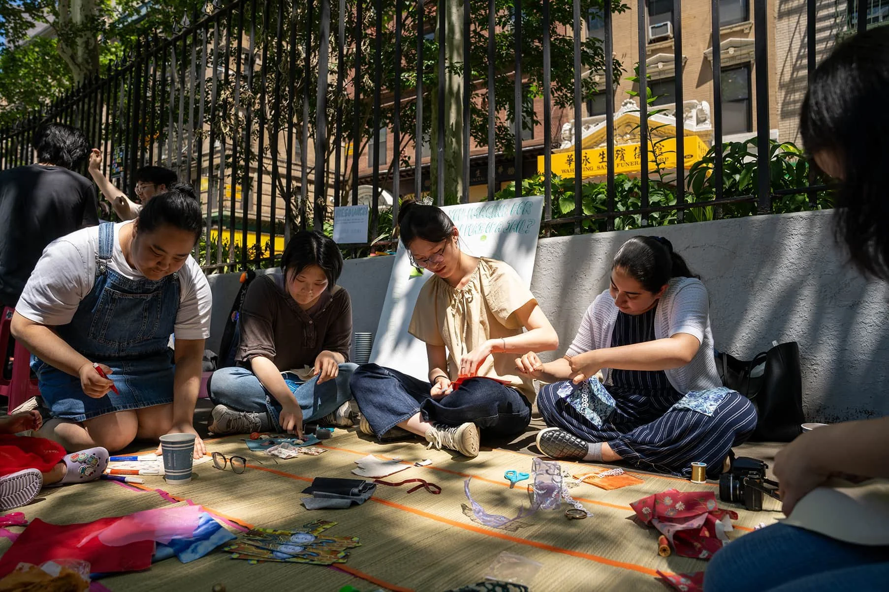 A group of women sitting on a mat outdoors, working on crafts with various supplies scattered around.
