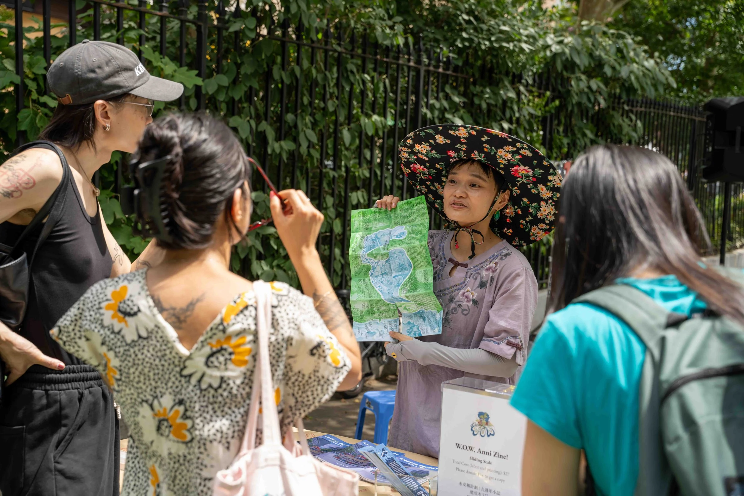 A woman with a large floral hat holding a map, speaking to a group of four women, with two women wearing glasses and one wearing a helmet, outdoors near a black metal fence and green foliage.