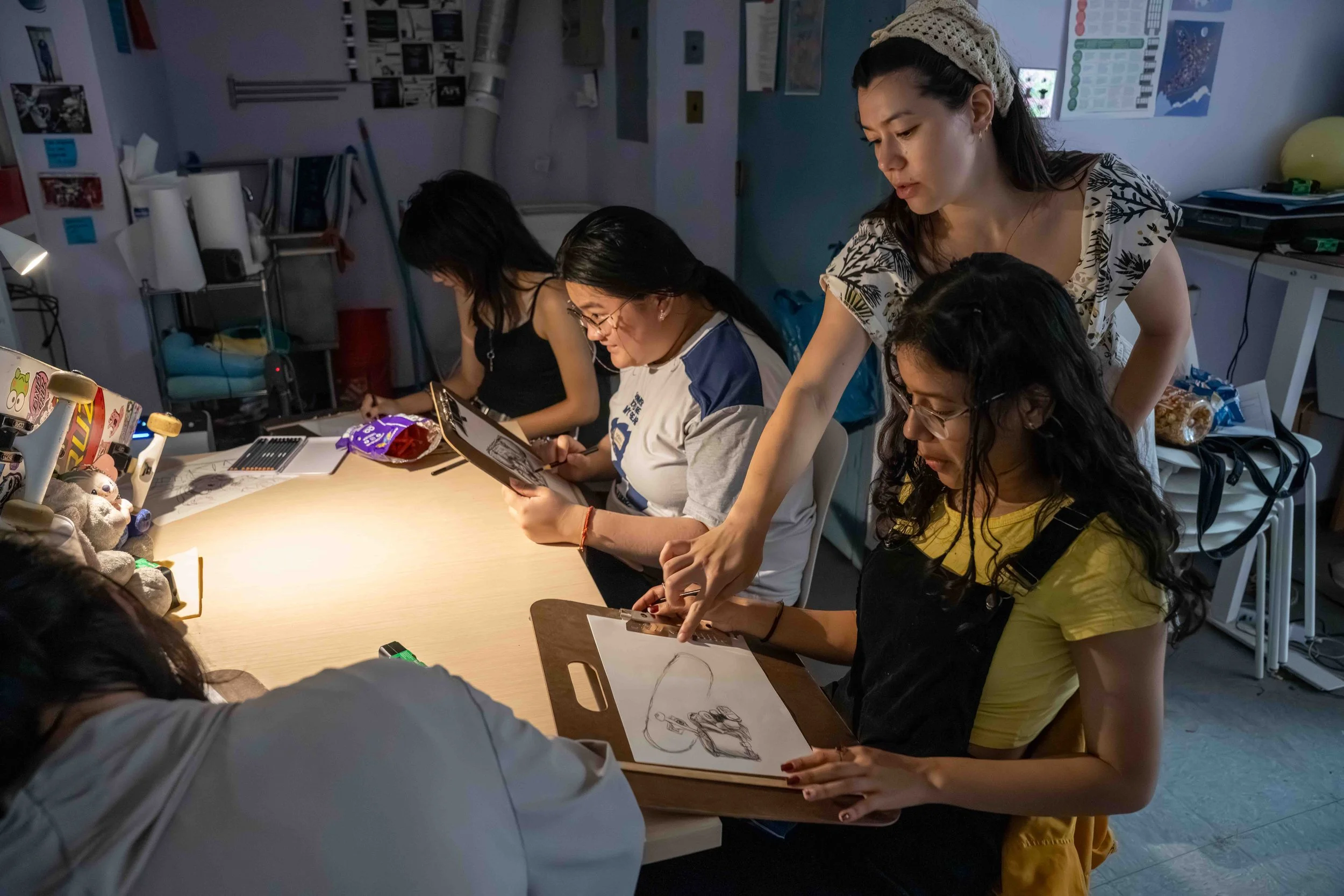 Group of women working on creative projects at a table in a well-lit room, with art supplies and drawings.