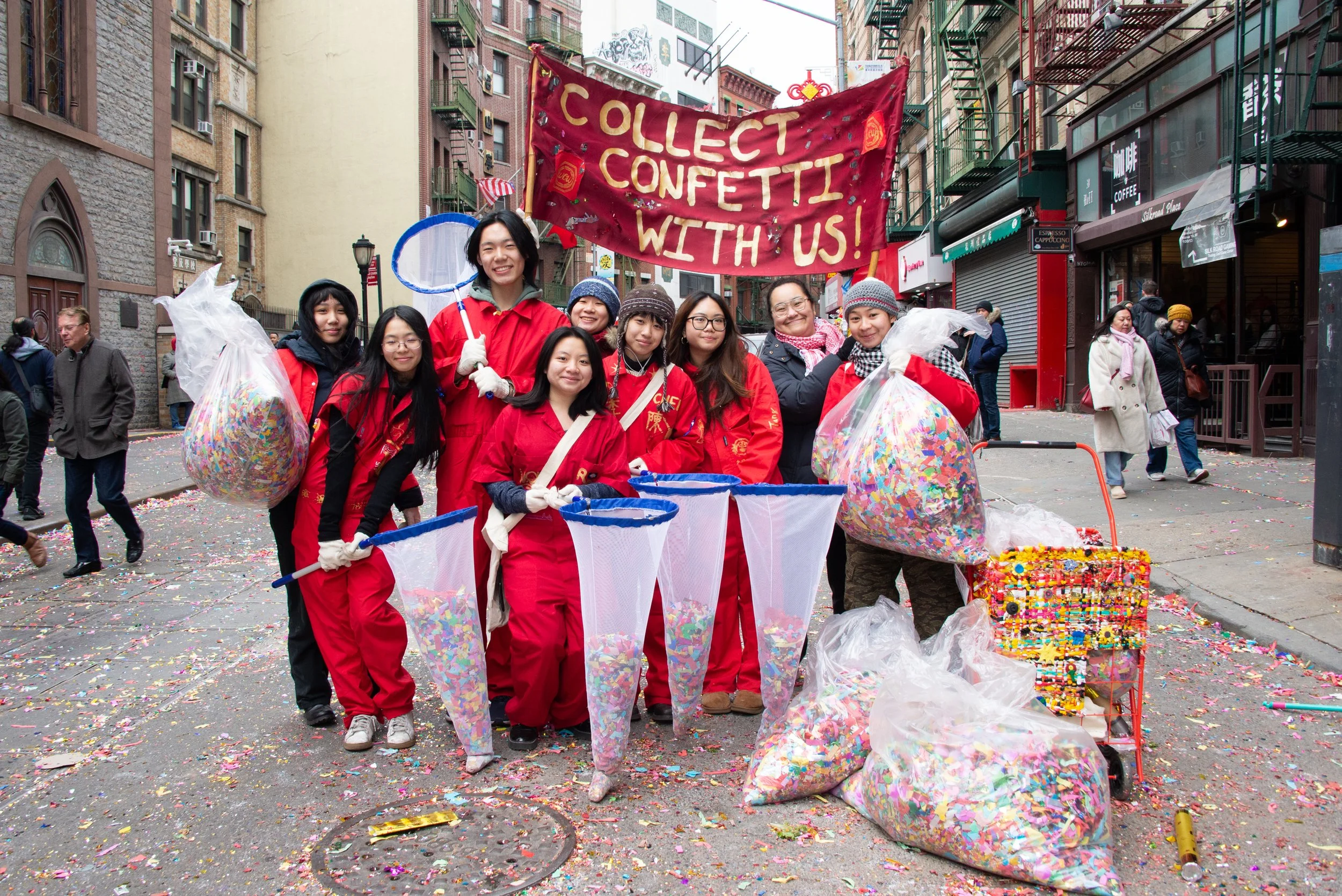 Group of people in red uniforms collecting confetti on a city street with a red banner that reads 'Collect Confetti With Us!'