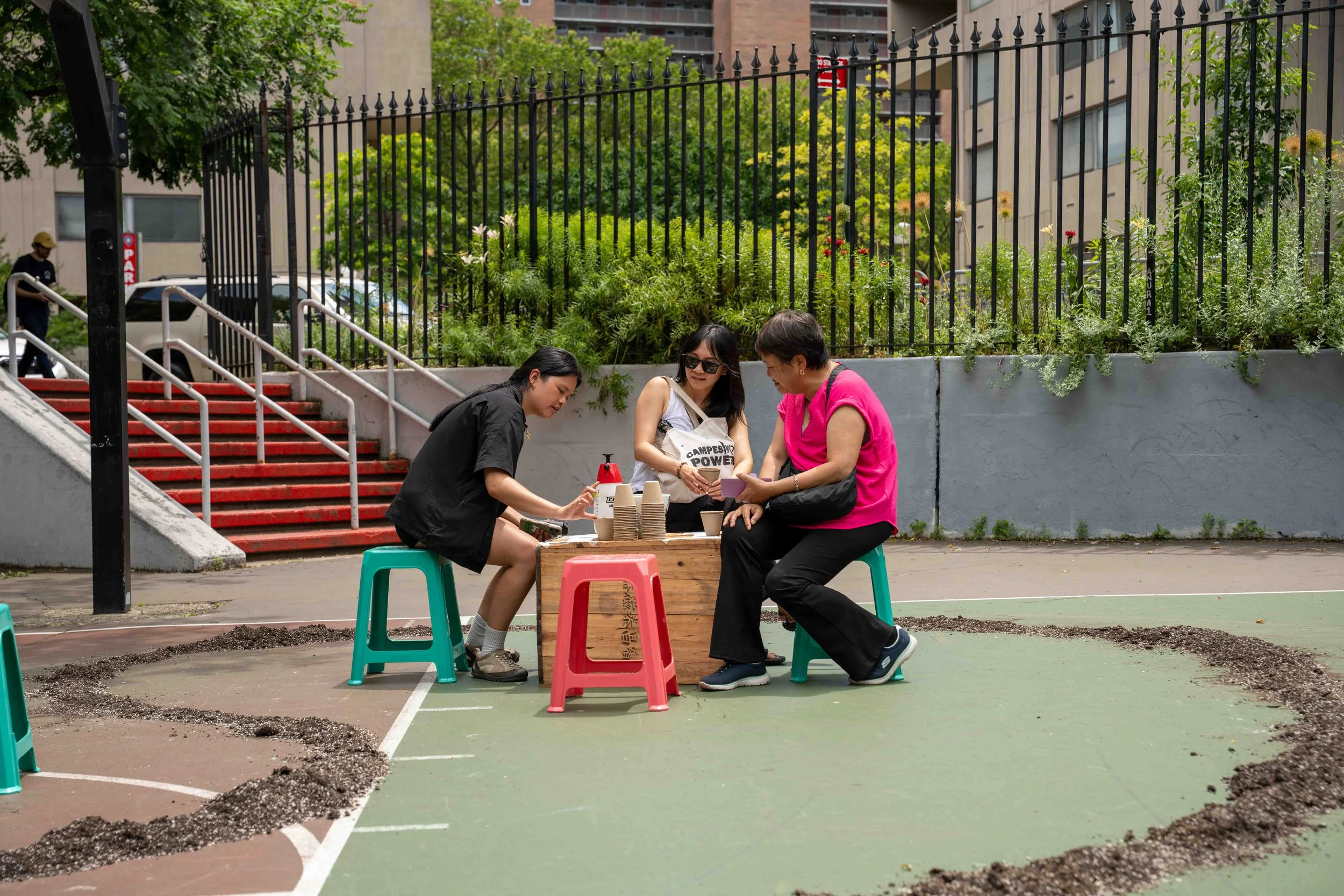 Three women sitting on colorful stools at a small table outdoors, with a basketball court and stairs with red treads in the background. They appear to be engaged in a conversation, surrounded by greenery and urban buildings.