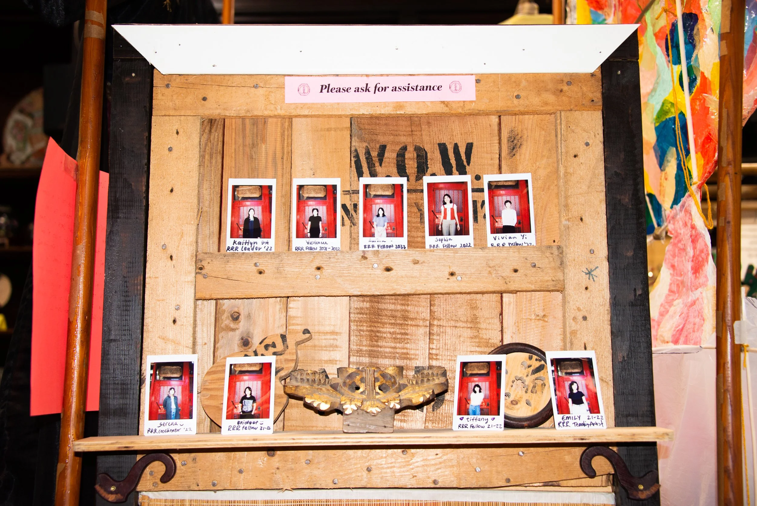 Polaroid photos of people pinned on a wooden display with a sign above that says, 'Please ask for assistance.'