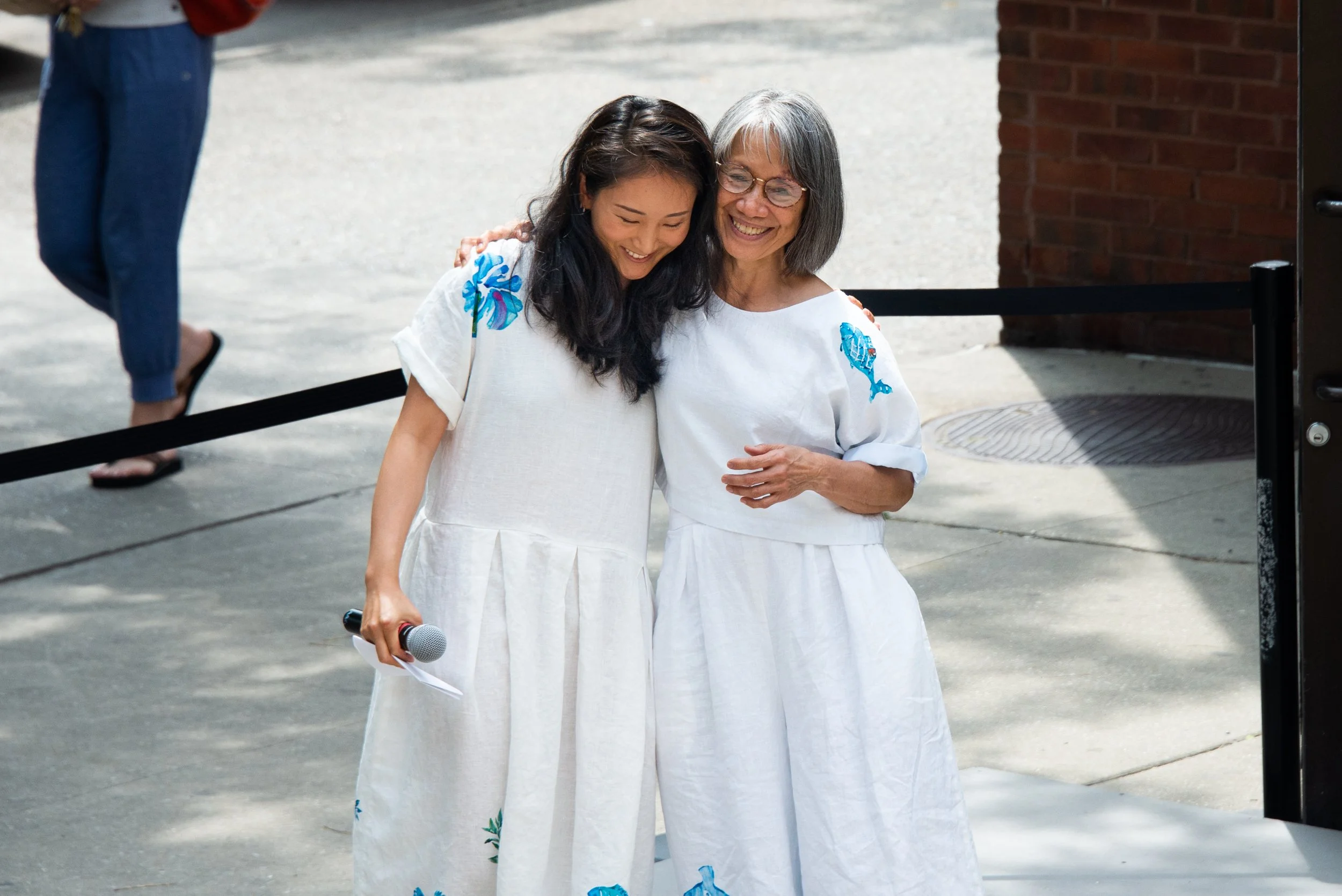 Two women dressed in white, smiling and hugging each other, one holding a microphone and paper, outside near a brick building.