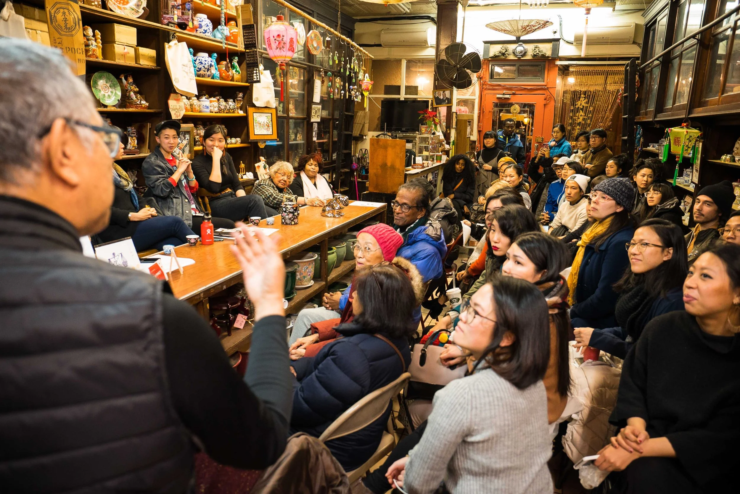 A group of people seated in a cozy, eclectic room decorated with Asian art and lighting, attentively listening to a person speaking at the front.