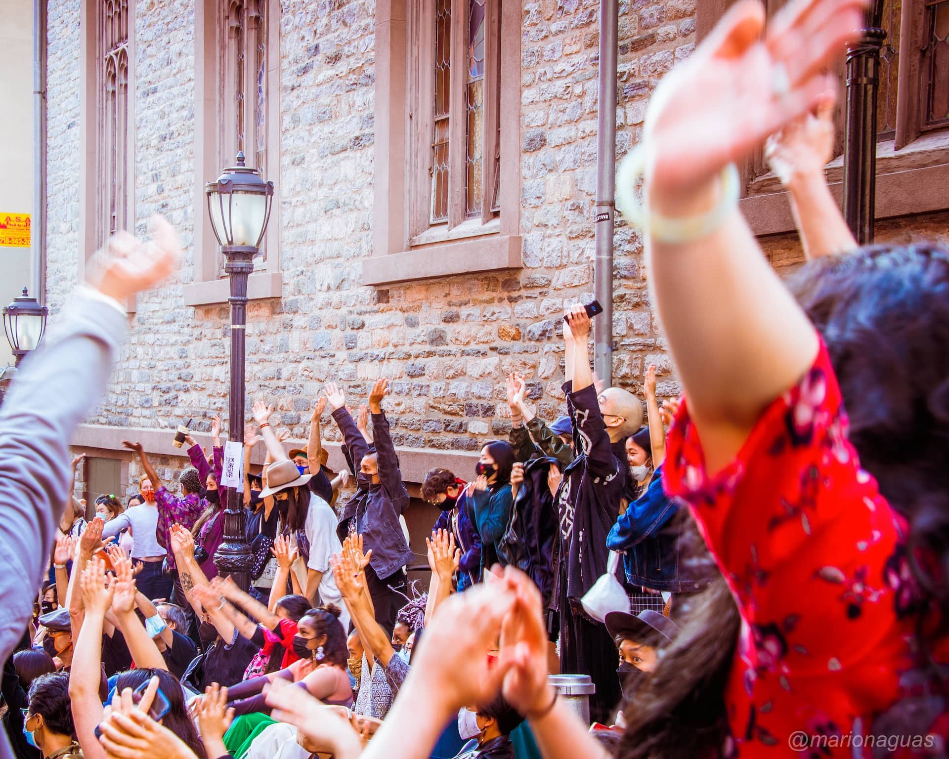A large group of people gathered outdoors in front of a brick building, raising their hands in the air during a protest or demonstration.