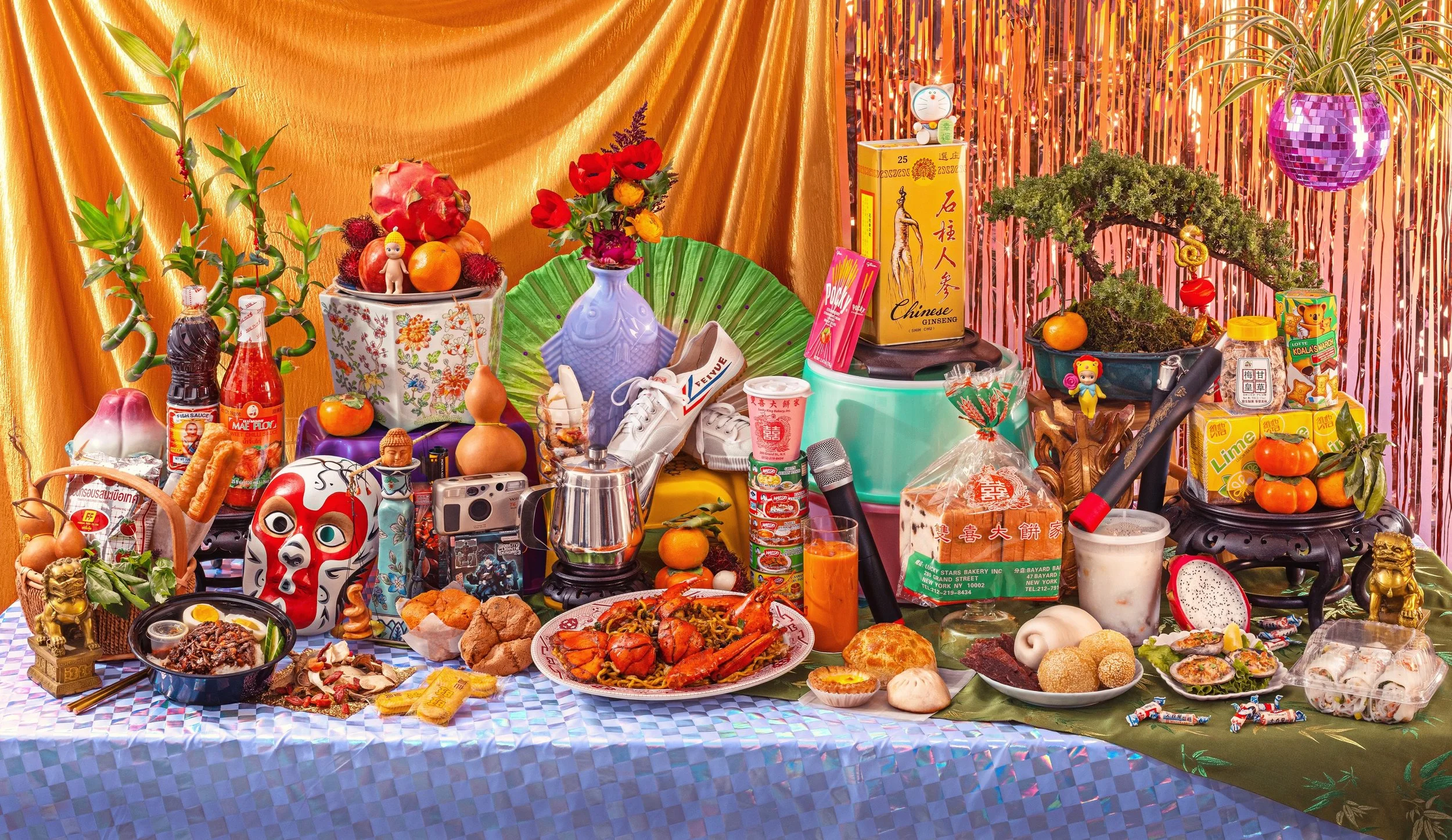 A table filled with various Asian foods and decorations, featuring fruits, snacks, drinks, and traditional ornaments on a colorful backdrop with curtains and plants.