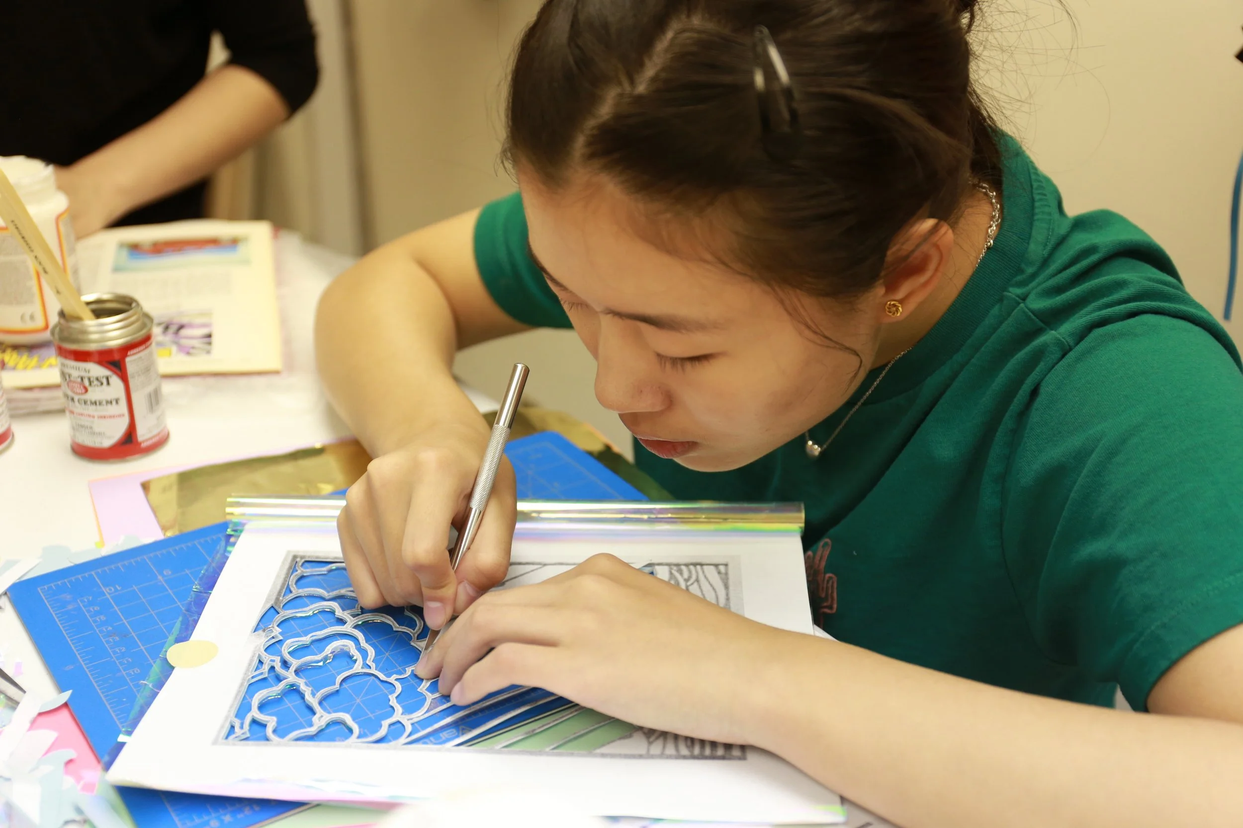 A young woman with dark hair and a hair clip, wearing a green shirt, is carefully cutting out a paper design with a craft knife at a desk surrounded by craft supplies and papers.