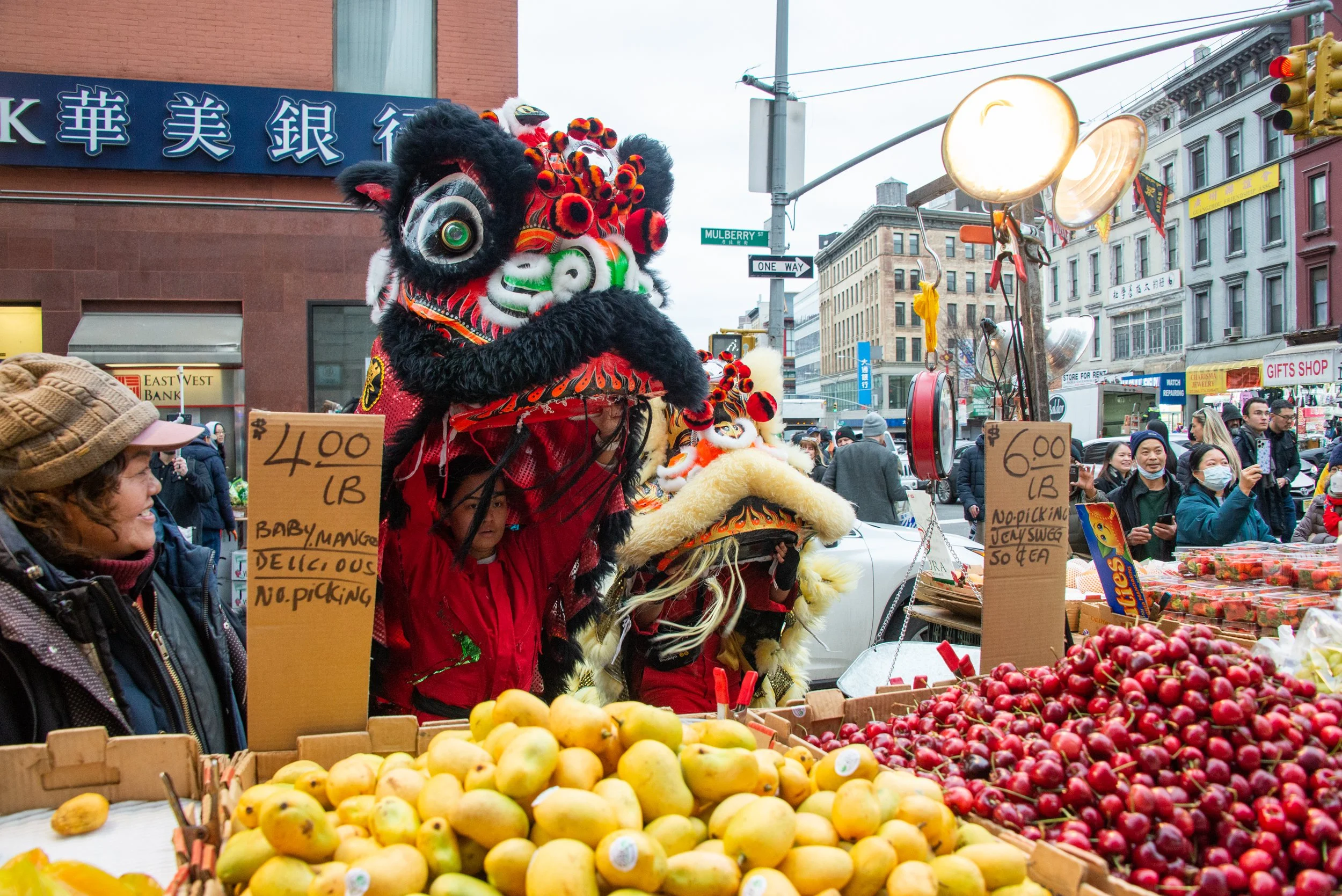 A street market scene with a person holding up a lion dance head for a lion costume, surrounded by fruit stalls with apples and pears, and various vendors and people in the background.
