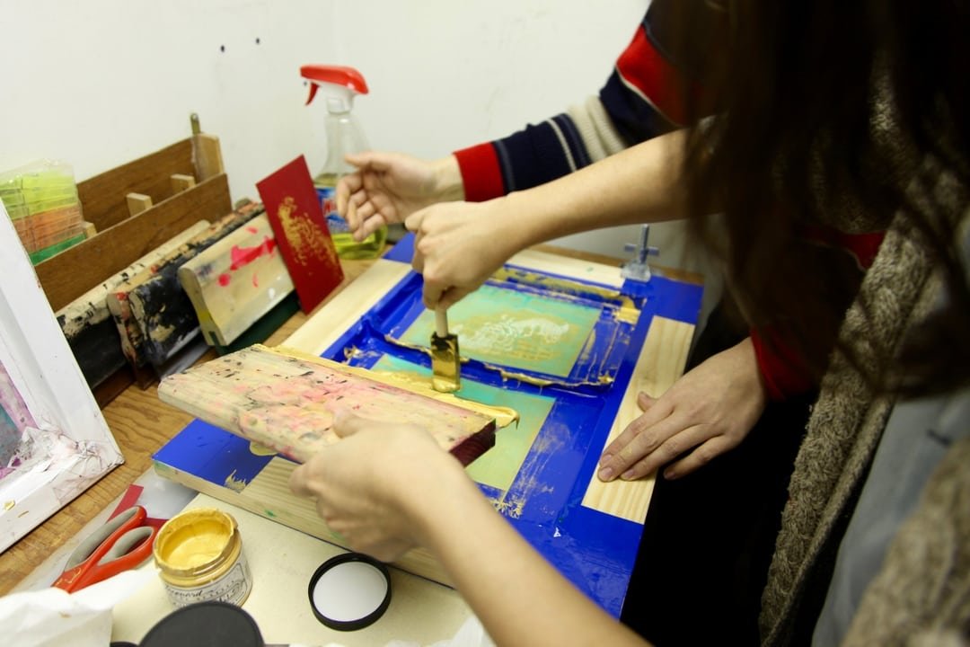 Two people using a brayer and woodblock printing on paper at a craft desk, with printing supplies around.