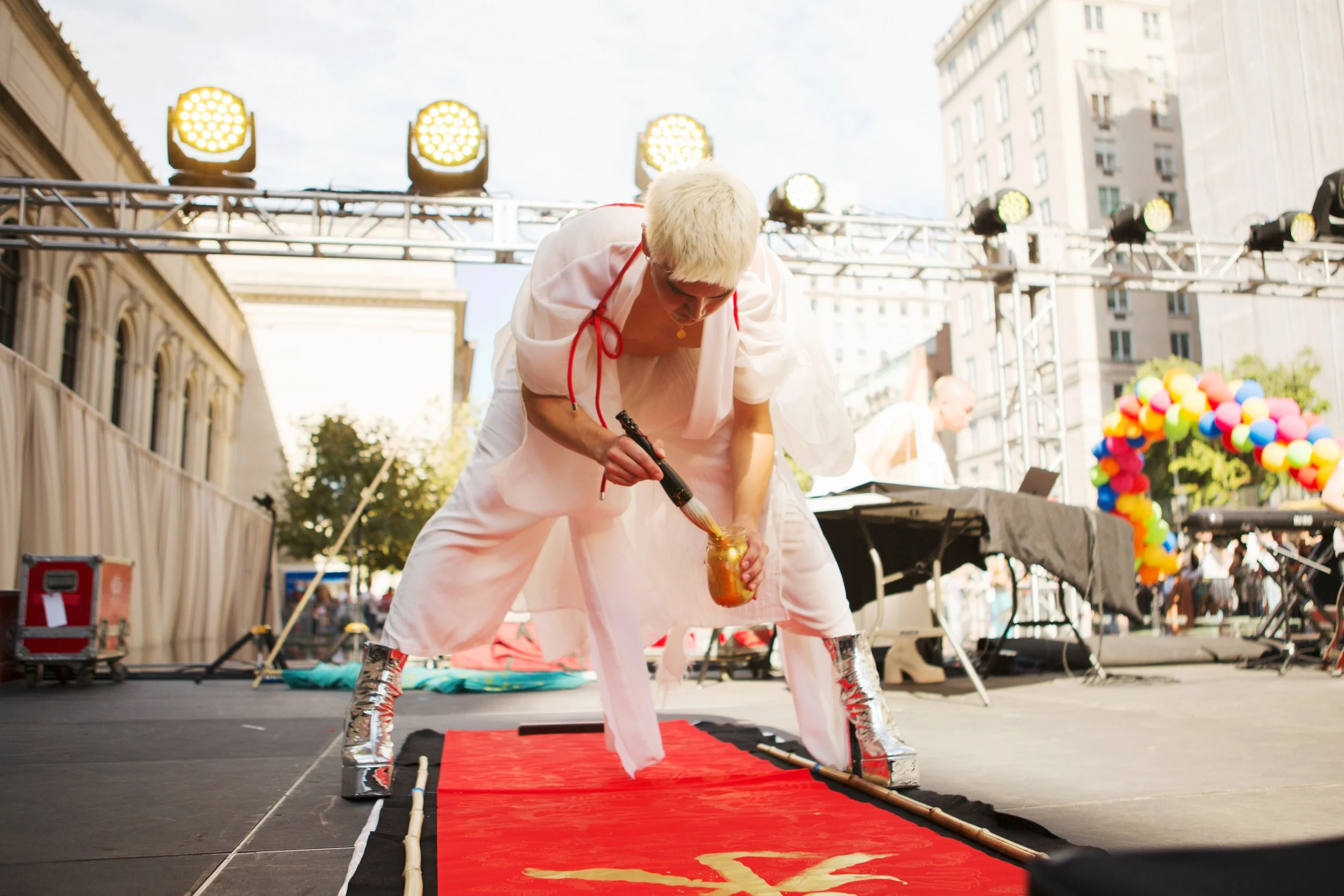 Person with short blond hair, dressed in white, painting on a red carpet with a brush during an outdoor event on a city street. Stage lights and a colorful balloon arch are visible in the background.