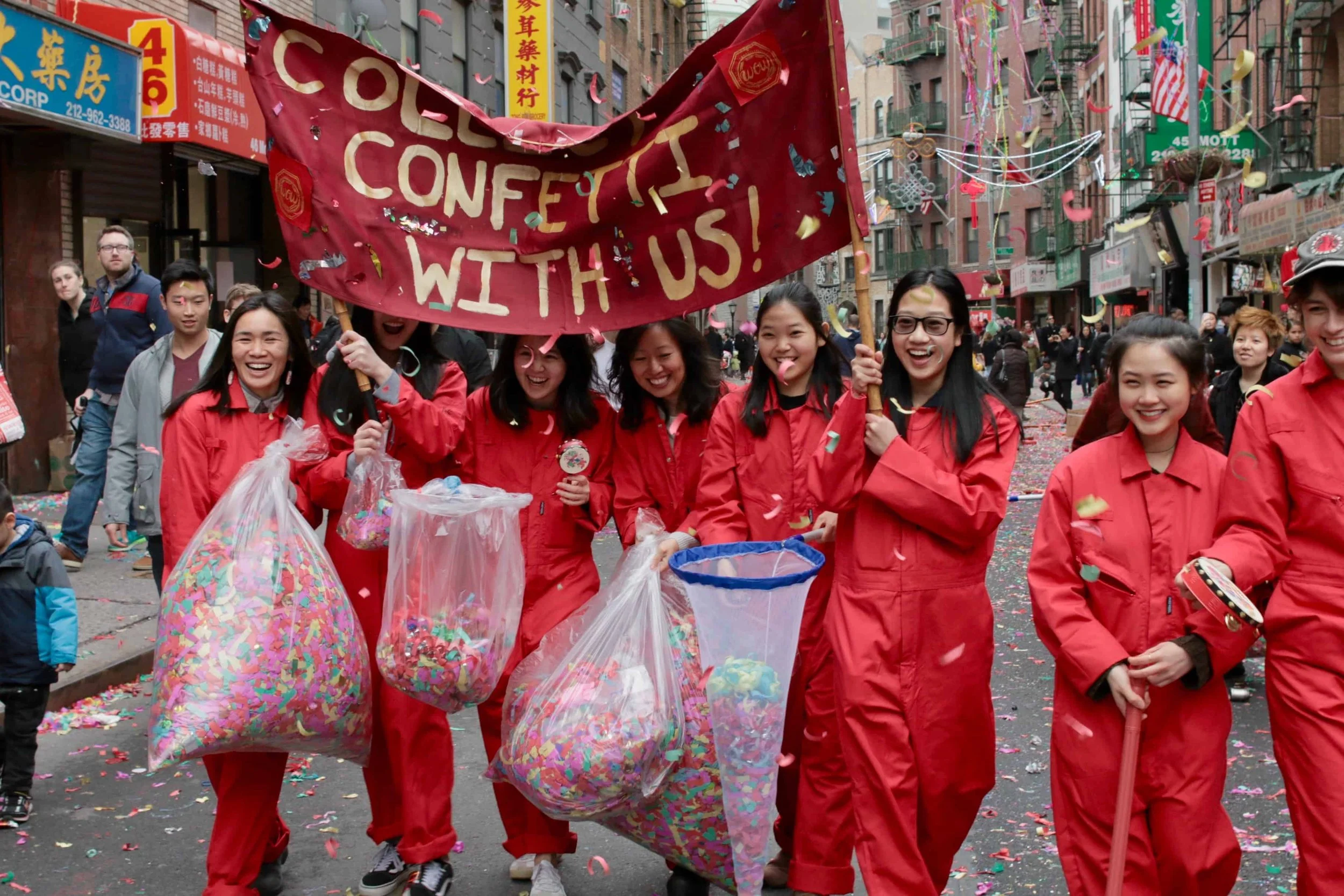 A group of women wearing red uniforms celebrating in a street parade with confetti, holding bags of confetti, and a banner that reads 'Celebrate with us!'