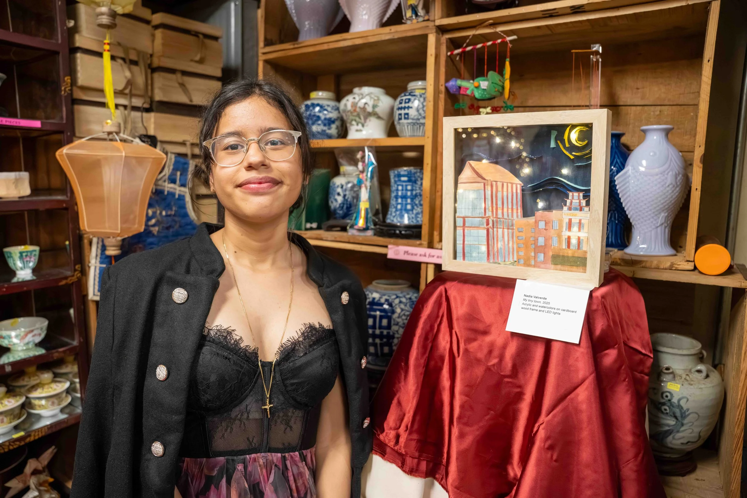 Young woman with glasses and a black lace top, standing in front of a display of pottery and artwork at an art exhibit.