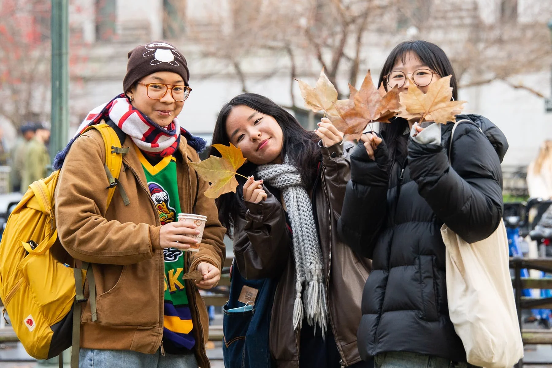 Three young women with backpacks standing outdoors in autumn, holding fallen leaves, smiling at the camera.