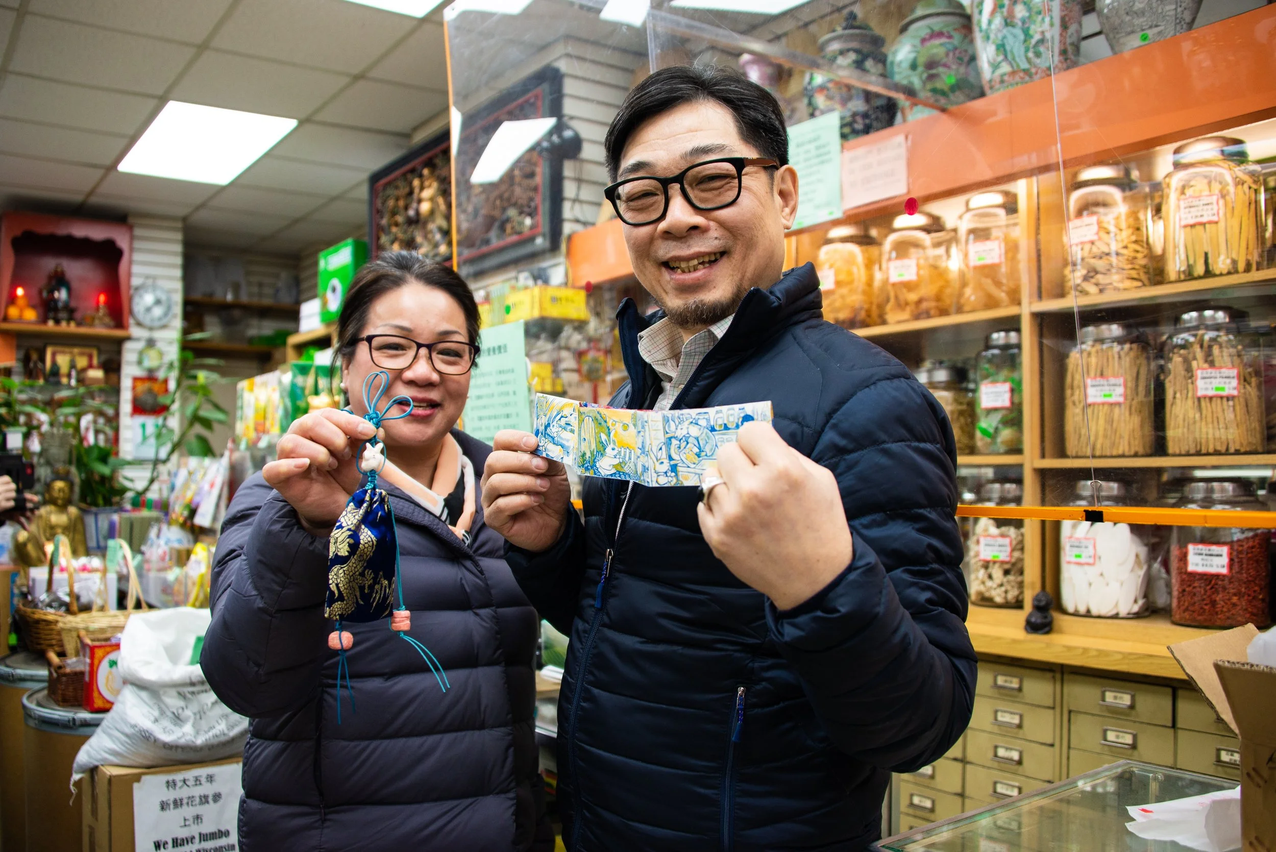 Two people in a store holding traditional Chinese New Year ornaments and paper money, smiling for the camera.