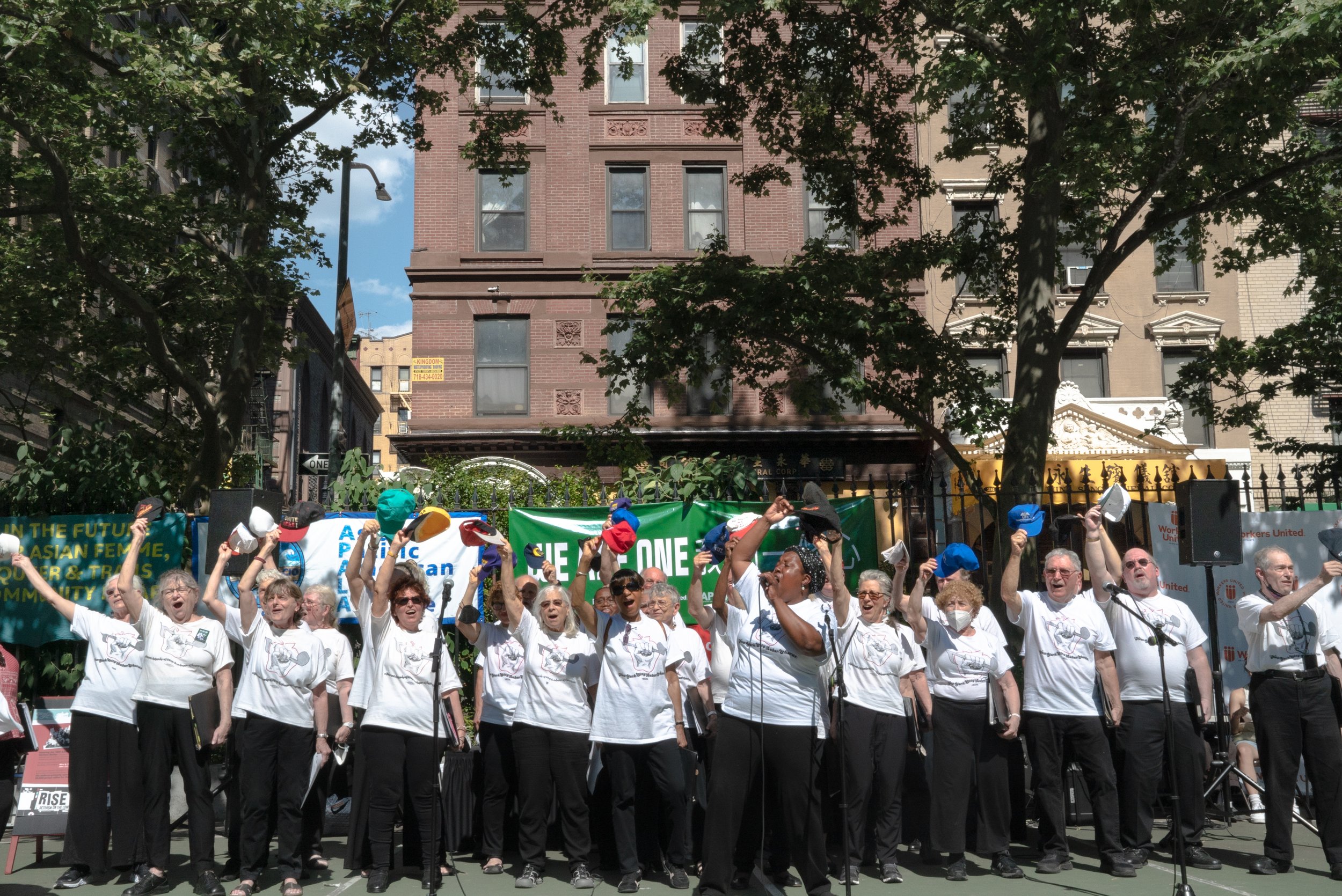 A group of diverse elderly people on a stage outdoors, waving hats in the air, singing or cheering, with banners and trees in the background.