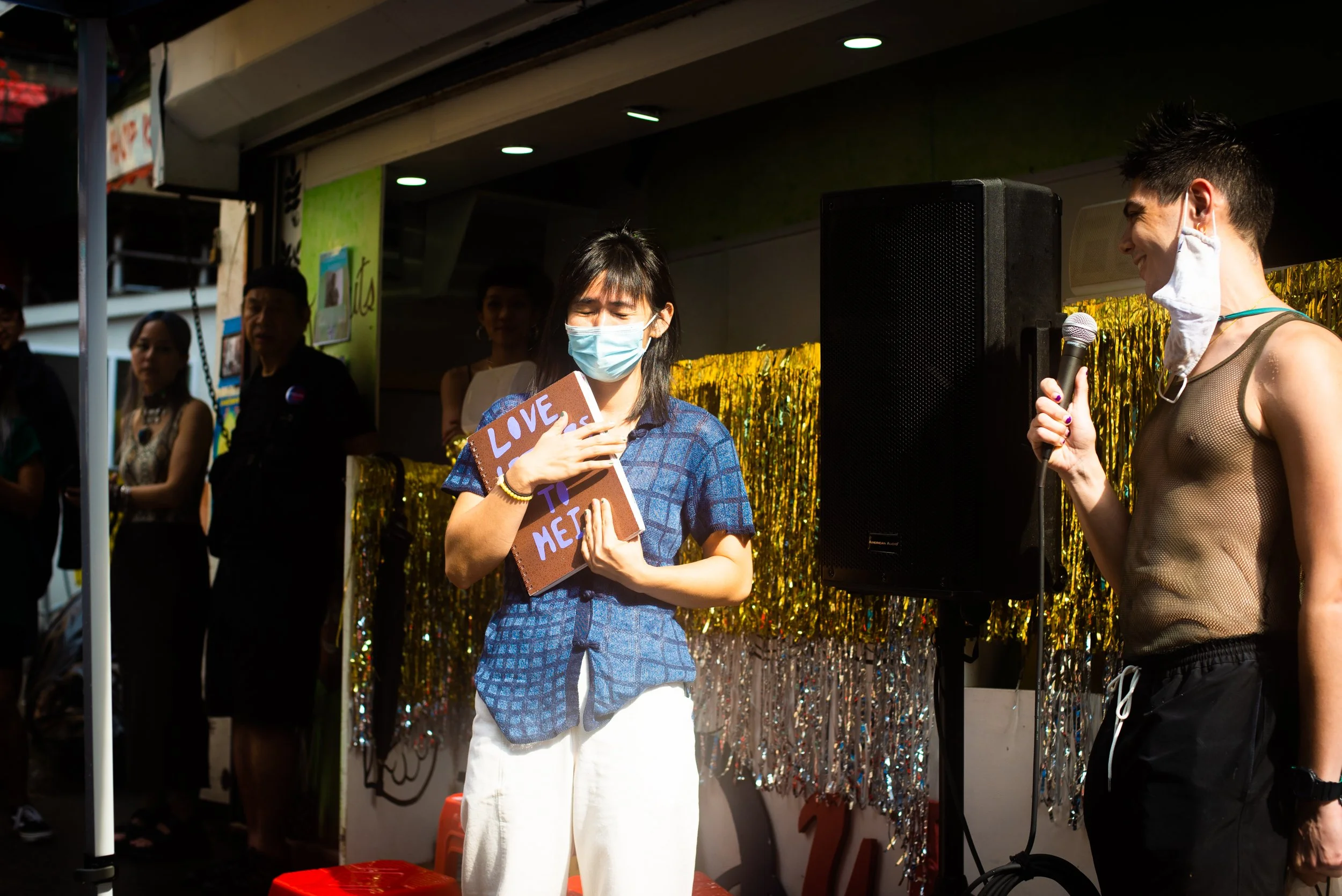 A person on stage at an event, holding a sign that says 'Love to Me,' while wearing a face mask. Another person with a microphone and an unbuttoned sleeveless top stands nearby with a mask around their neck. Several people are in the background, with a shiny gold and silver backdrop.