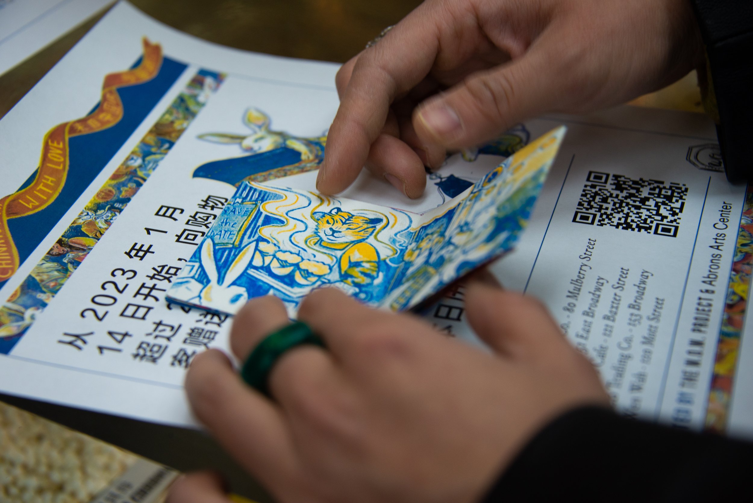 Person peeling back a flyer or ticket with colorful illustrations, including a tiger, rabbit, and a ribbon, with text in Chinese and English, and a QR code.