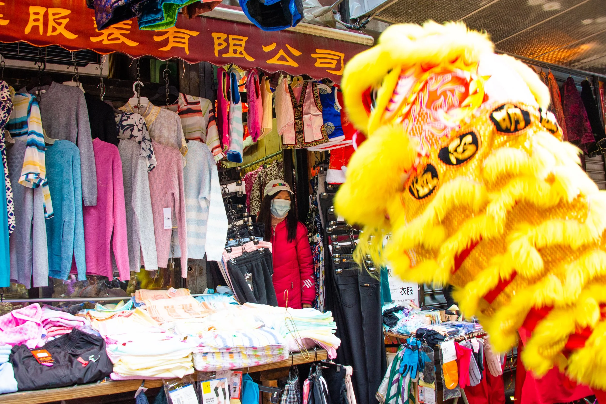 A street market stall selling colorful sweaters, shirts, and accessories with a person wearing a face mask standing behind the stall, and a blurry yellow lion costume in the foreground.