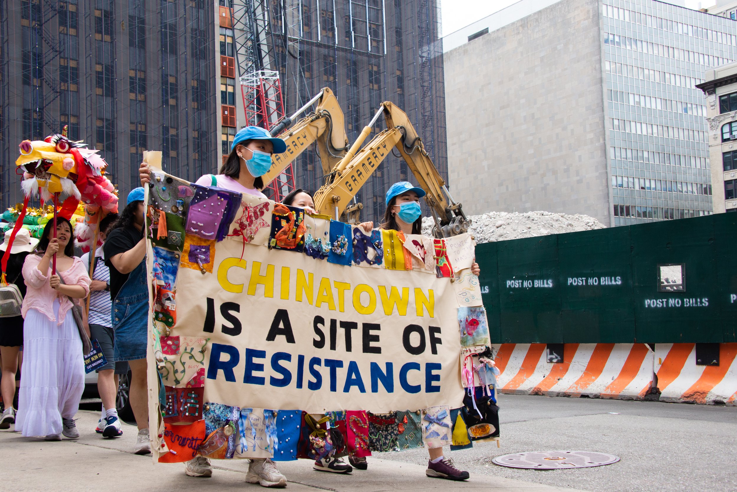 People participating in a protest march, holding a large handmade sign that reads 'Chinatown is a site of resistance.' Some individuals are wearing face masks and colorful clothing, including dragon costumes. A construction site with machinery and a green barrier is visible in the background.