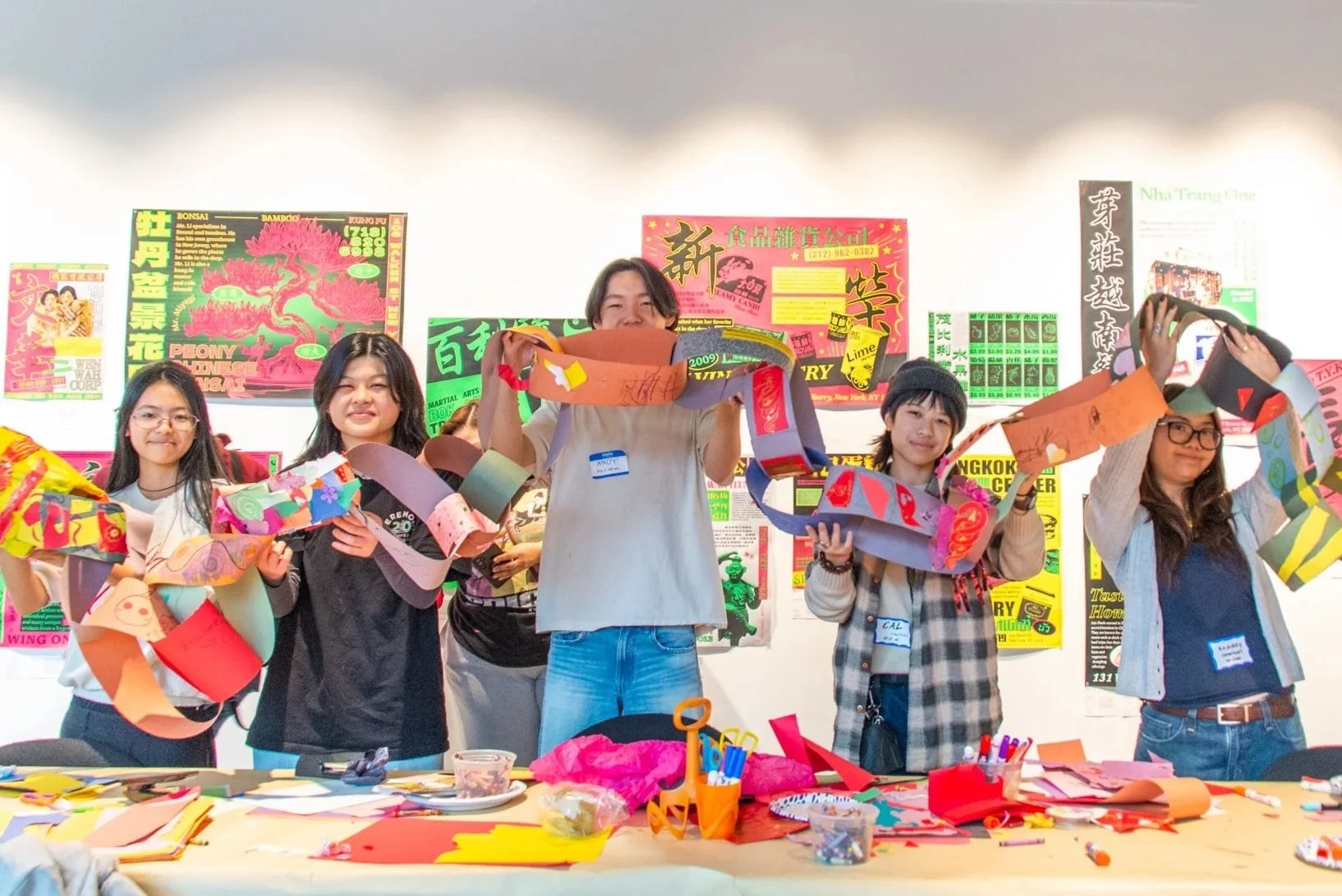 Group of people making colorful paper chain decorations in a room with posters on the wall.