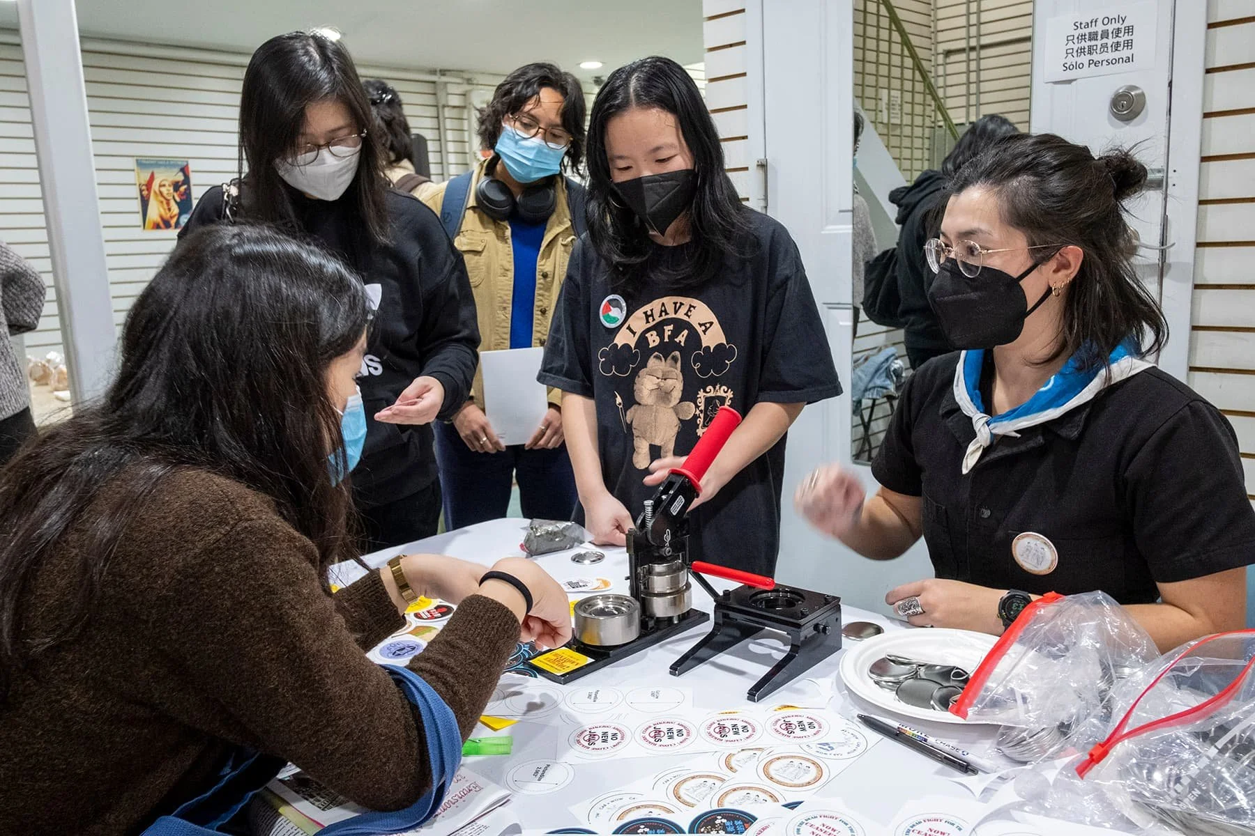 Group of women in masks gathered around a table with a heat press and stickers, working on a craft project.