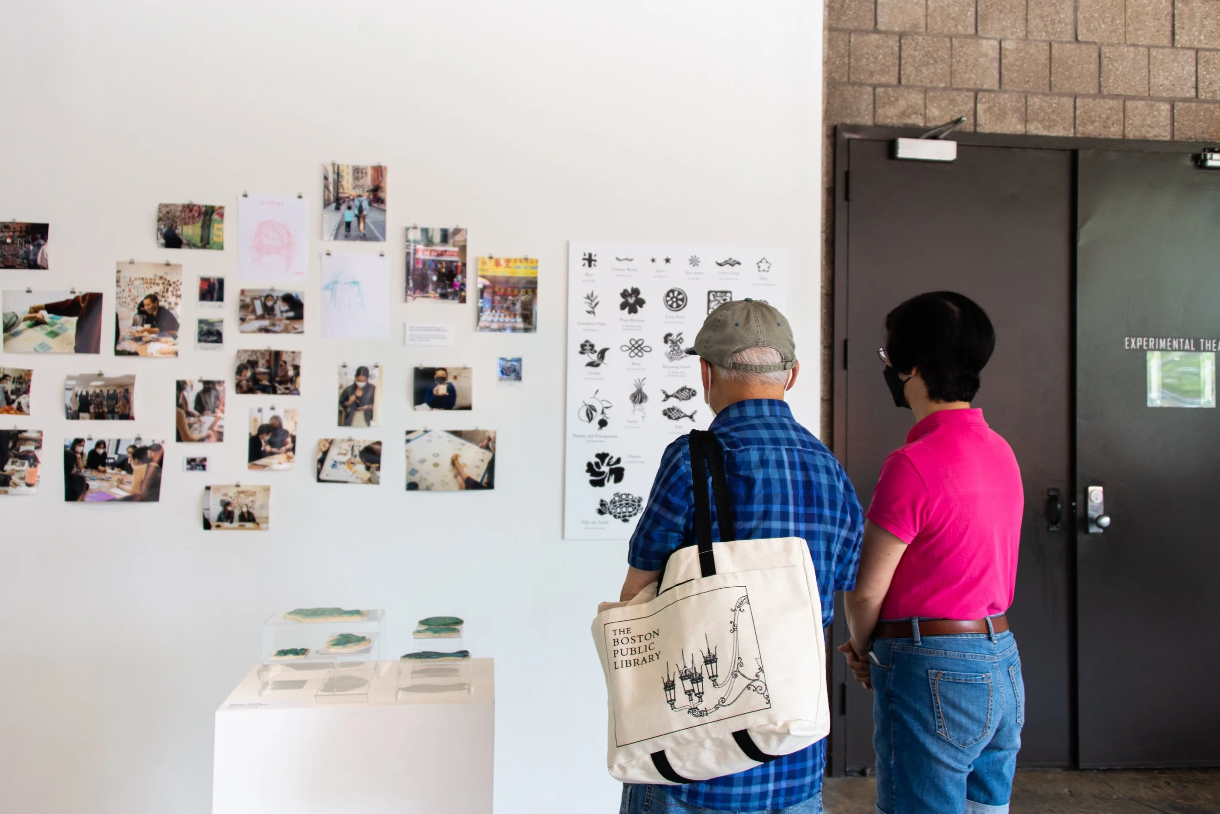 Two people observe artwork displays on a white wall at an art gallery. One wears a blue plaid shirt, a gray cap, and carries a tote bag labeled 'The Boston Public Library.' The other is dressed in a pink shirt and jeans, both with their backs to the camera.