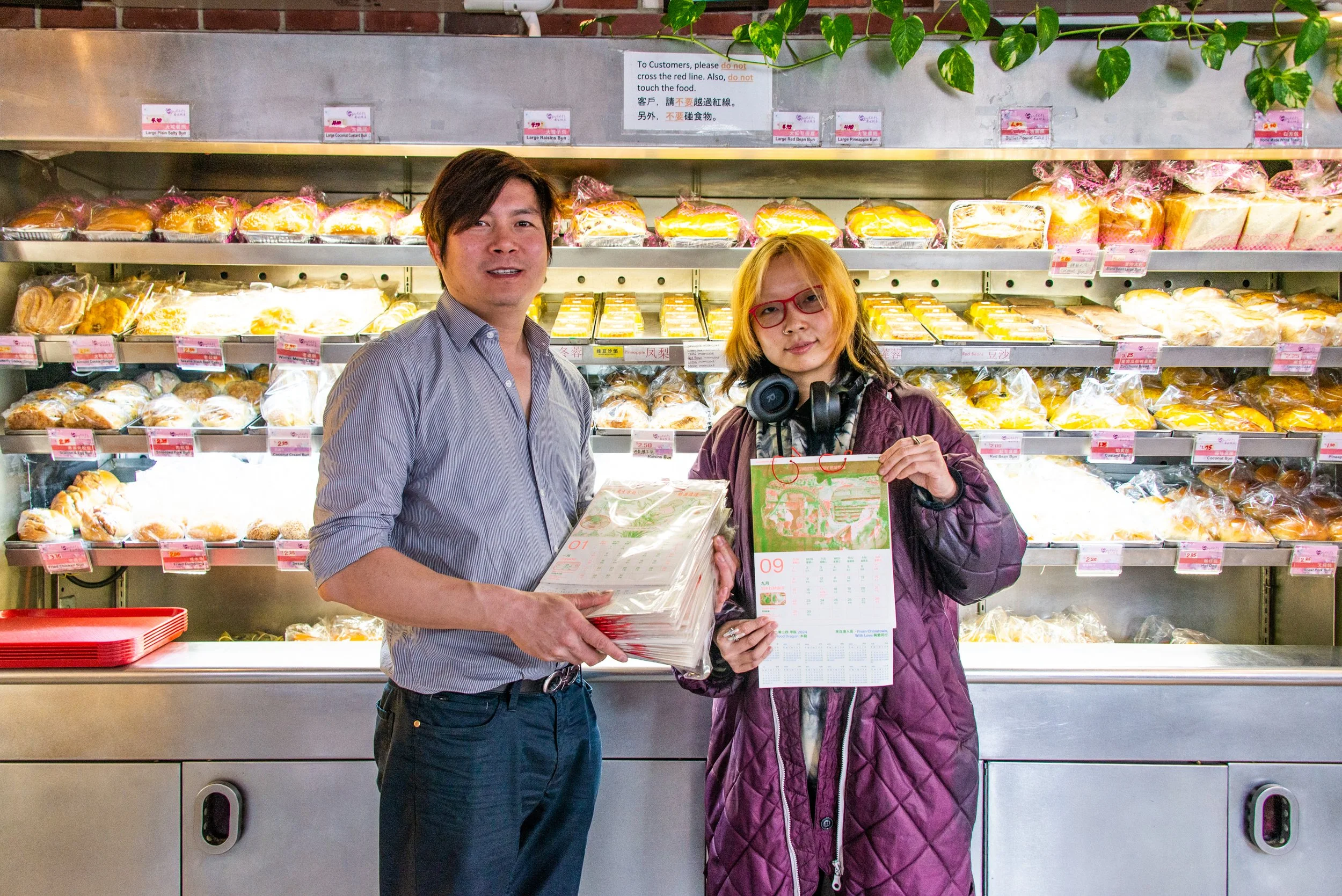 A man and a woman standing in front of bakery shelves holding a calendar and a stack of papers. The bakery shelves behind them are filled with packaged bread and baked goods.
