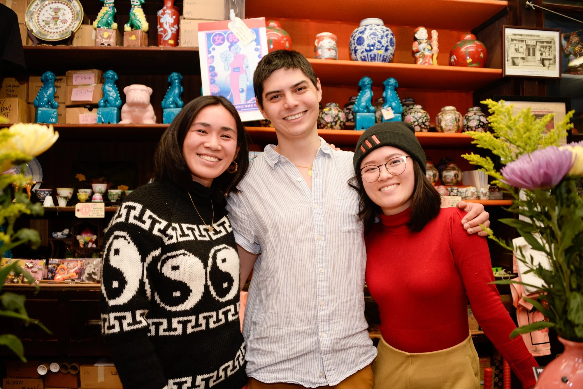 Three young adults smiling and standing close together in a colorful store with shelves filled with decorative pottery, figurines, and floral arrangements.