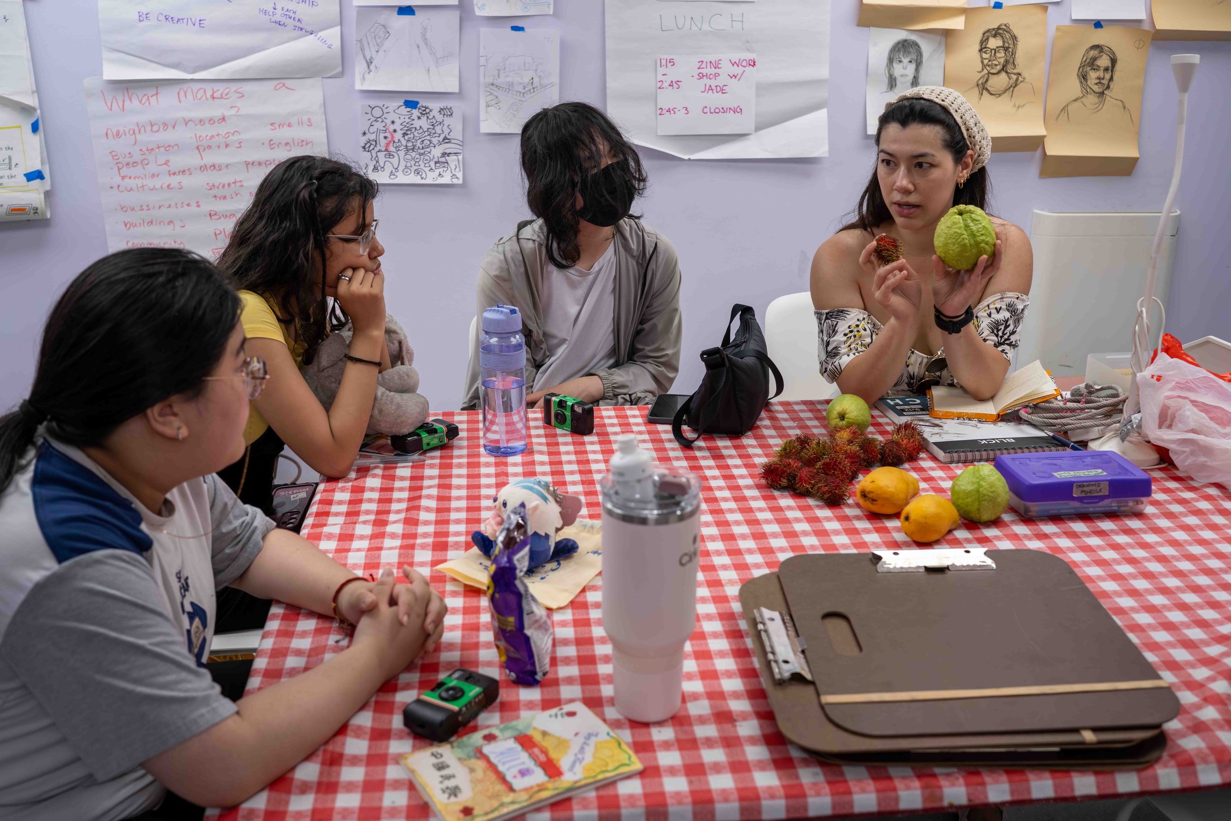 A group of five women sitting around a red and white checkered table, engaging in a discussion about fruits and tropical plants. The woman on the far right is holding a large green fruit and a smaller red fruit, with a notebook and a pile of rambutans, a guava, and other fruits on the table. The background features drawings, notes, and sketches taped to the wall.