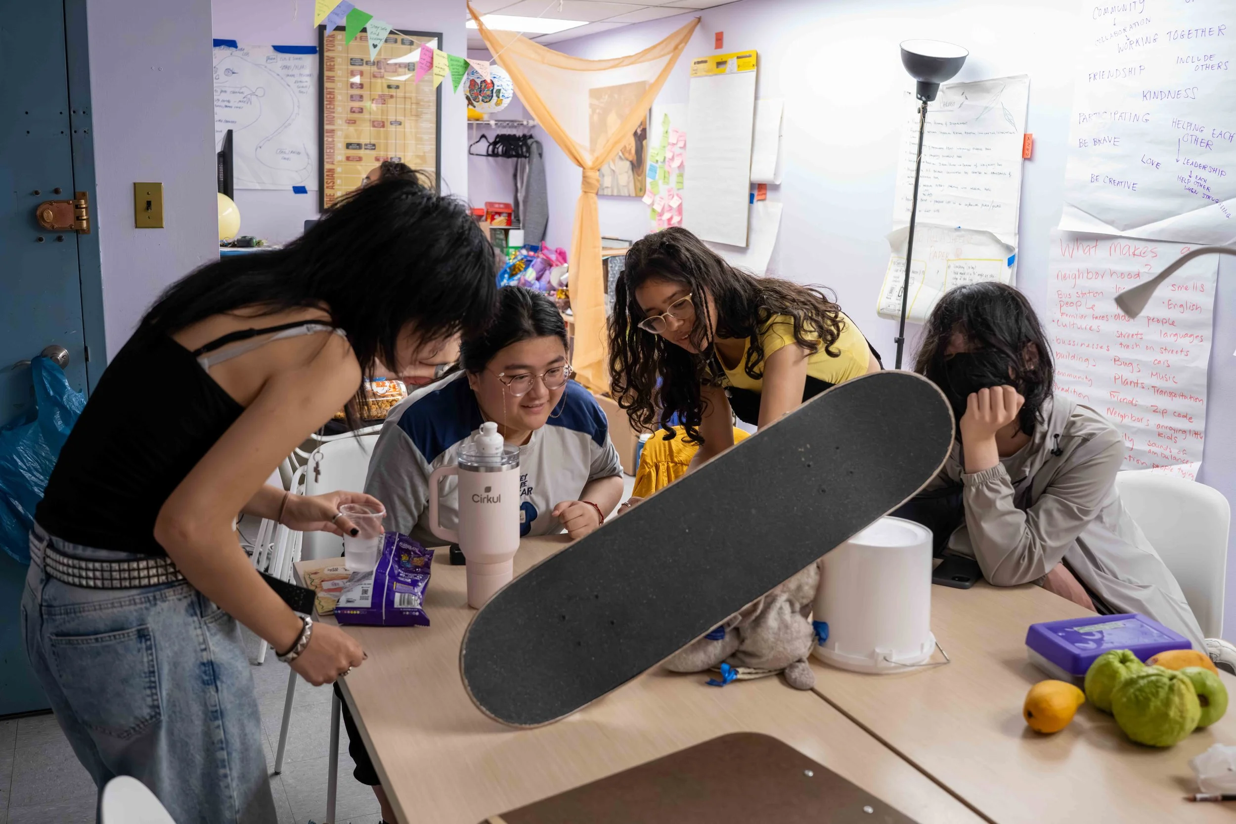 Five young women gathered around a table in a classroom, looking at something on the table, with posters and notes on the wall behind them, and various objects like fruit and a skateboard on the table.