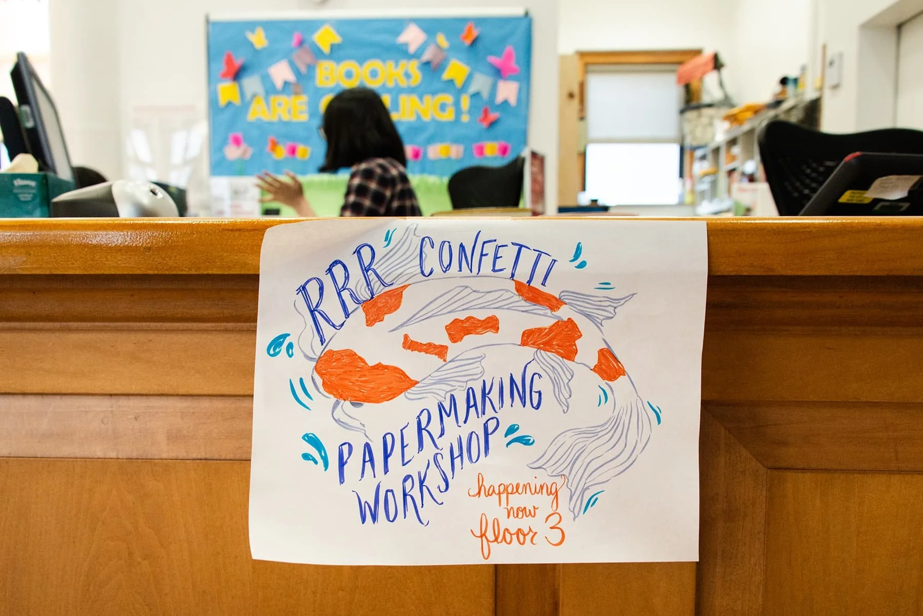 Colorful hand-drawn poster on a wooden counter announcing a paper-making workshop called 'RRR' Confetti, happening on the third floor, with a person working at a desk and a bulletin board that says 'Books Are Calling' in the background.