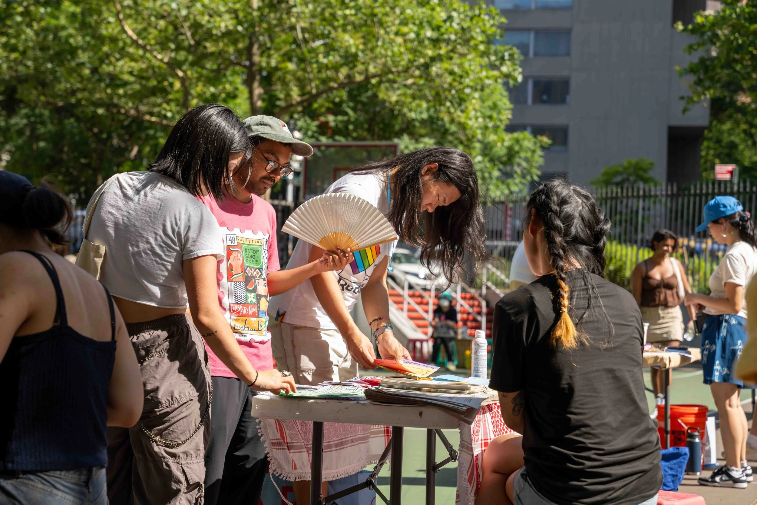 A group of people gathered at an outdoor table during daytime, with some browsing through papers, a person holding a colorful fan, and others engaged in conversation, in a park with trees and buildings in the background.
