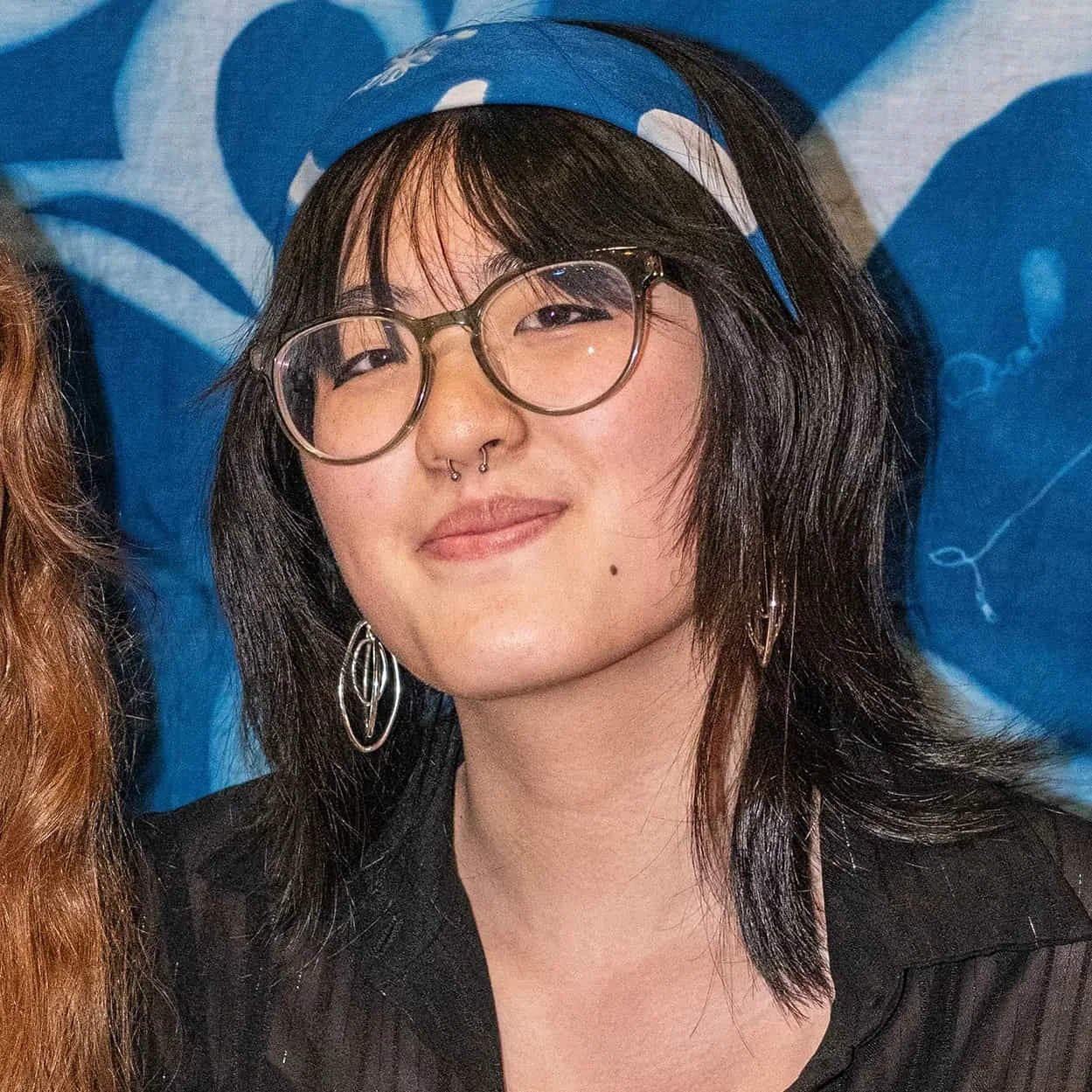 A young woman with glasses, septum piercing, and earrings, smiling in front of a blue background.