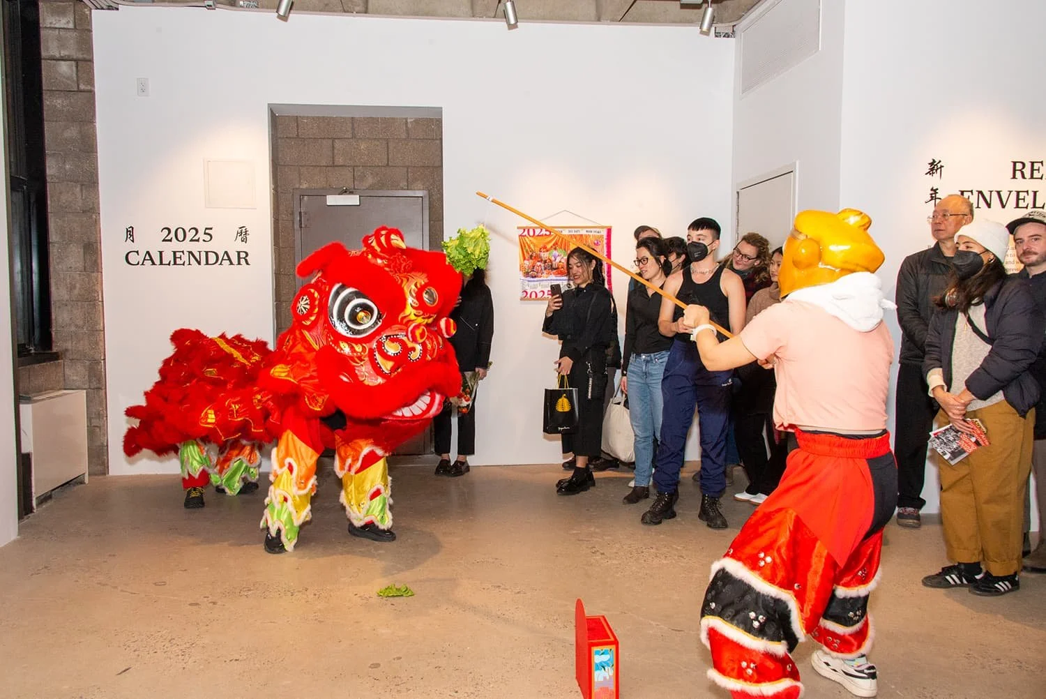People watching a traditional lion dance performance in an indoor space with a wall calendar and a poster. The lion costume is bright red with intricate gold and black detailing, and a performer with a lion head mask and red pants is holding a wooden stick, engaging with the lion. Audience members are standing, some holding cameras or smartphones, observing the performance.