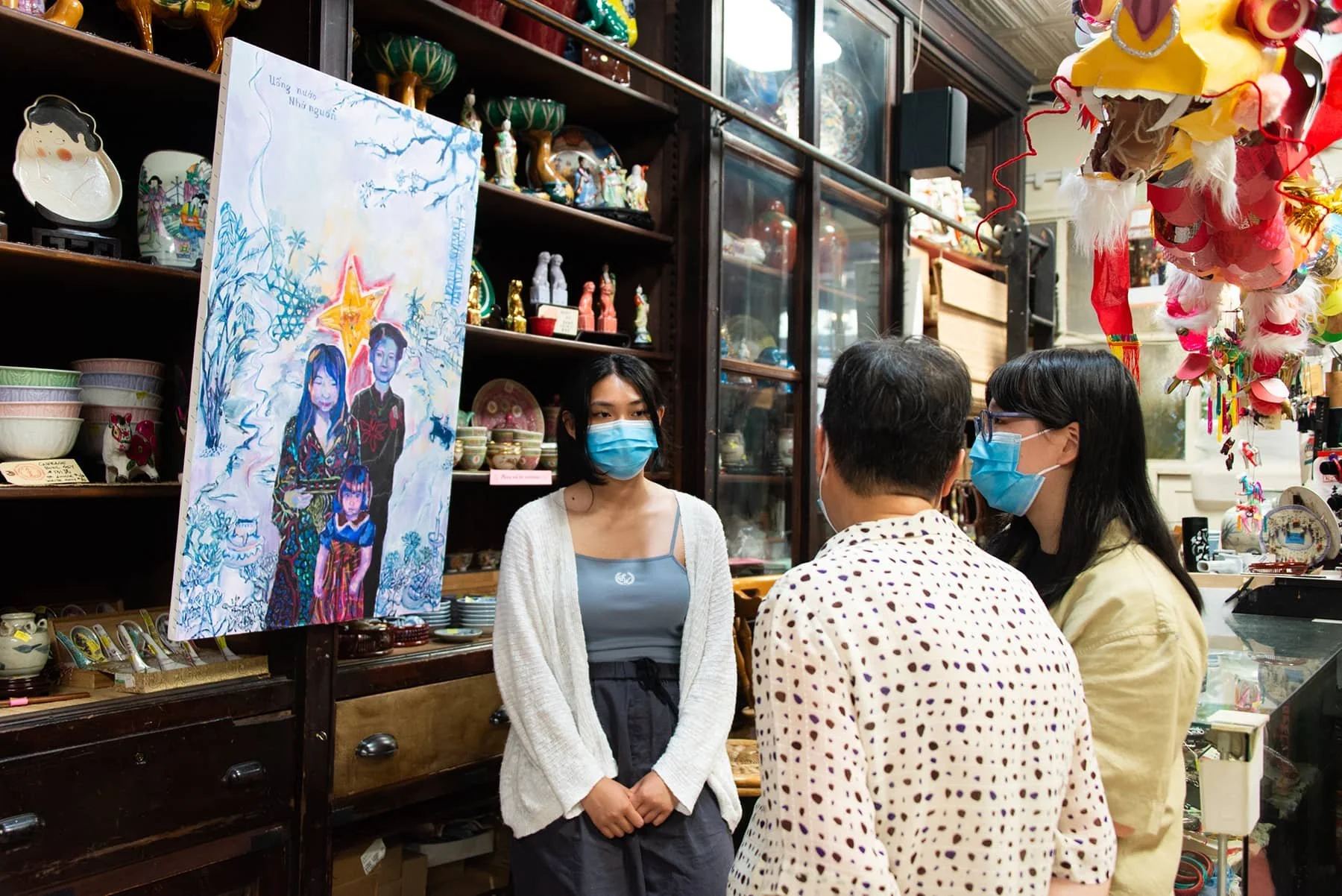 Three people in masks engaged in conversation inside a shop filled with ceramics and decorative items. A colorful artwork of a family hangs on the wall behind them.
