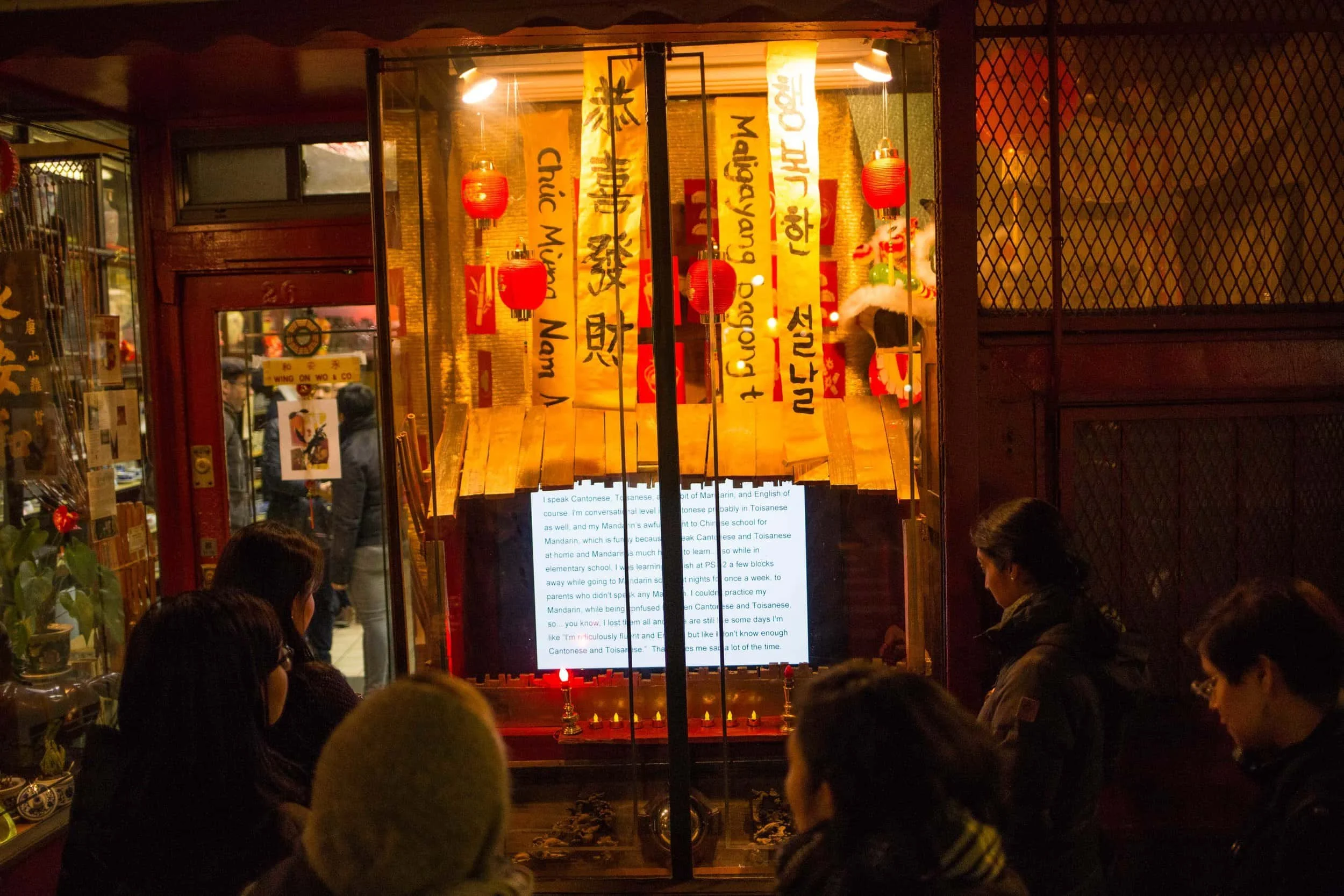 People gathered outside a storefront decorated for a celebration, with red lanterns and yellow banners with Chinese writing, and a digital display showing text.