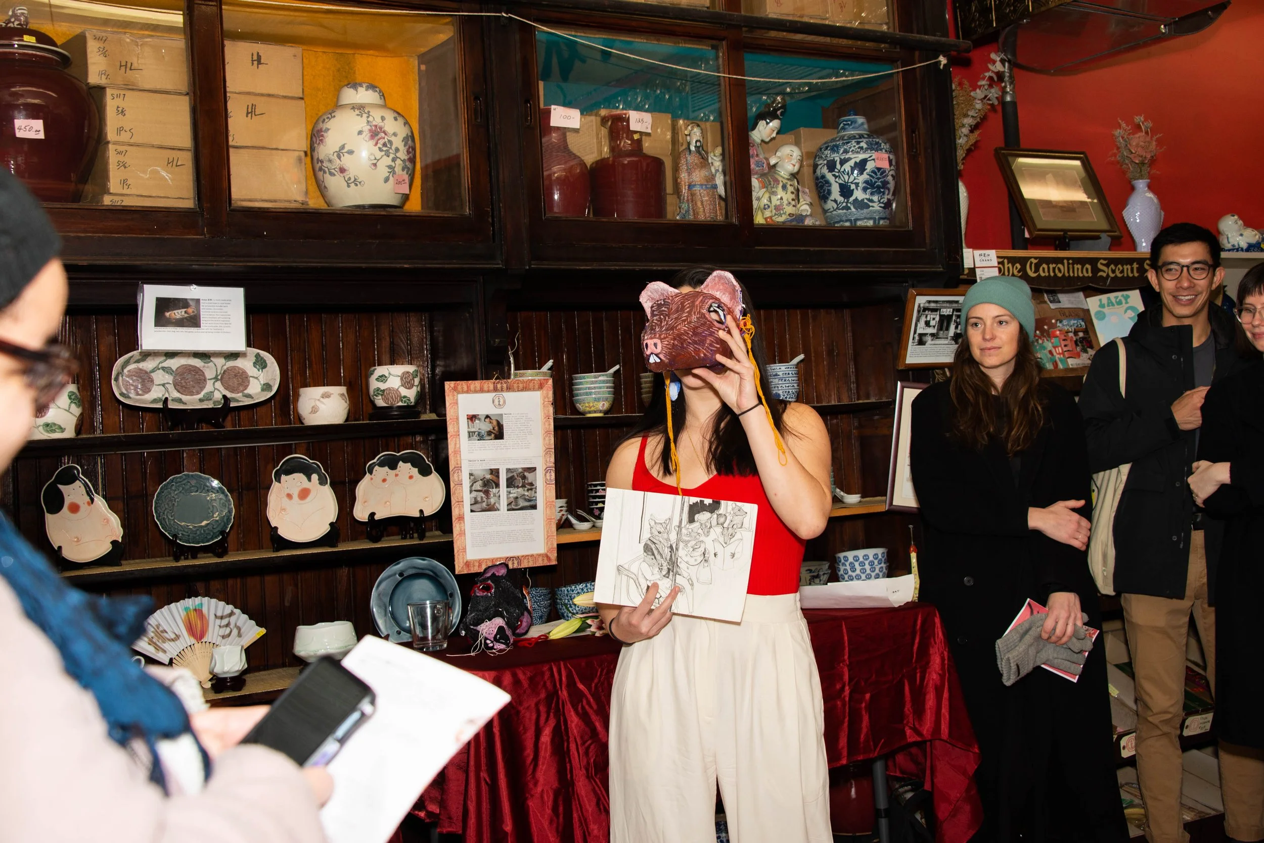 A group of people inside an antique shop, with one woman wearing a pig mask and holding a sketchbook, and others observing and taking photos.