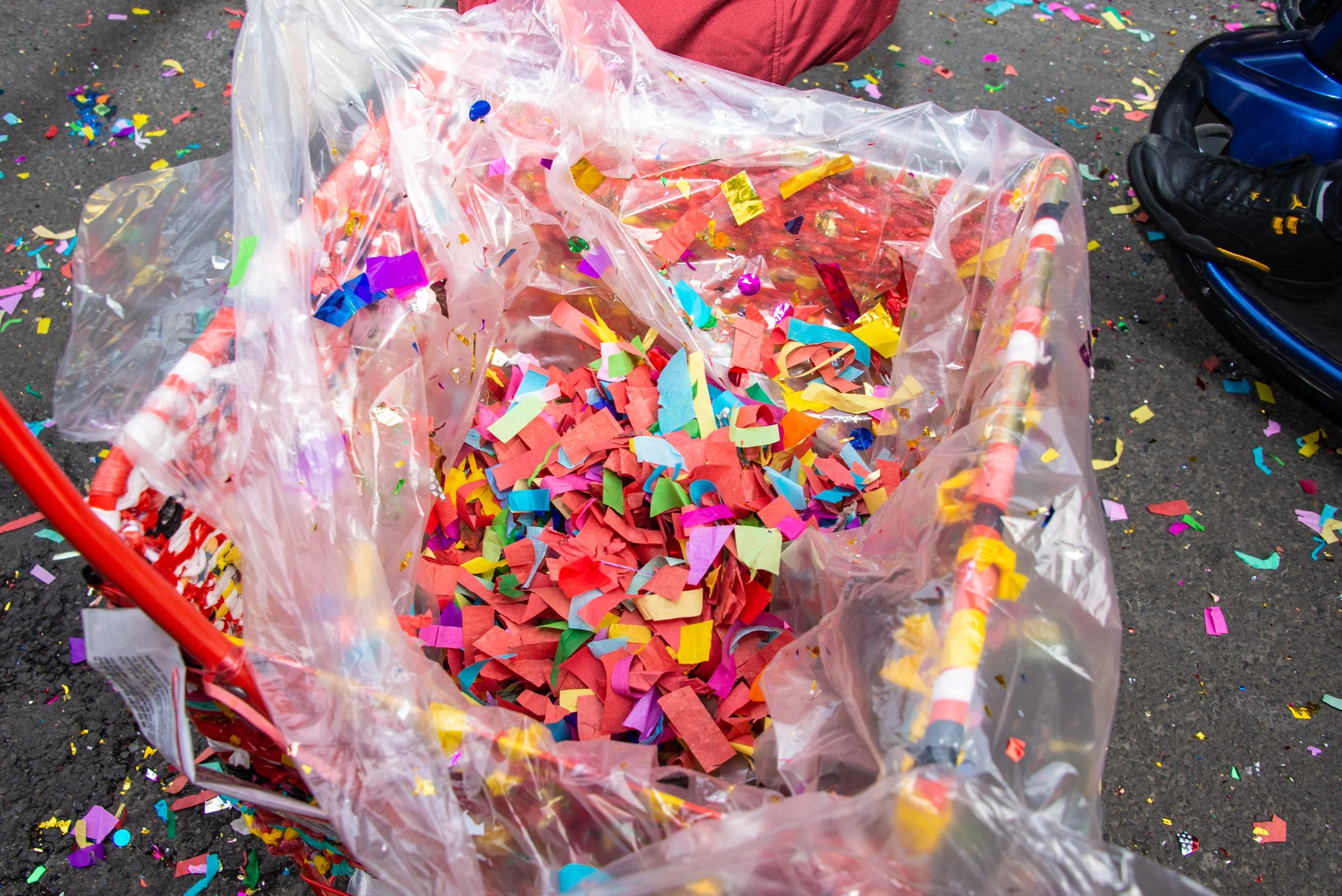 A large clear plastic bag filled with colorful confetti on an asphalt ground, with some confetti scattered around. Part of a person's leg in red pants and a sneaker are visible nearby.