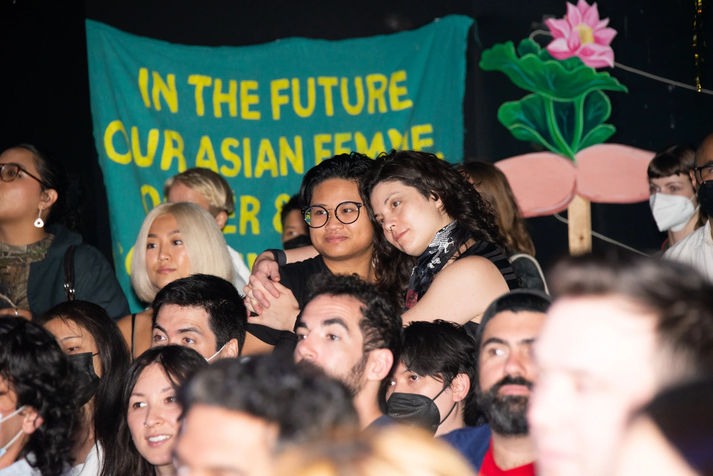 Group of diverse people at a public event, with some wearing masks, seated and watching attentively. A blue banner with yellow text in the background reads, 'In the future our Asian female,' and there are colorful lotus flower and plant illustrations.