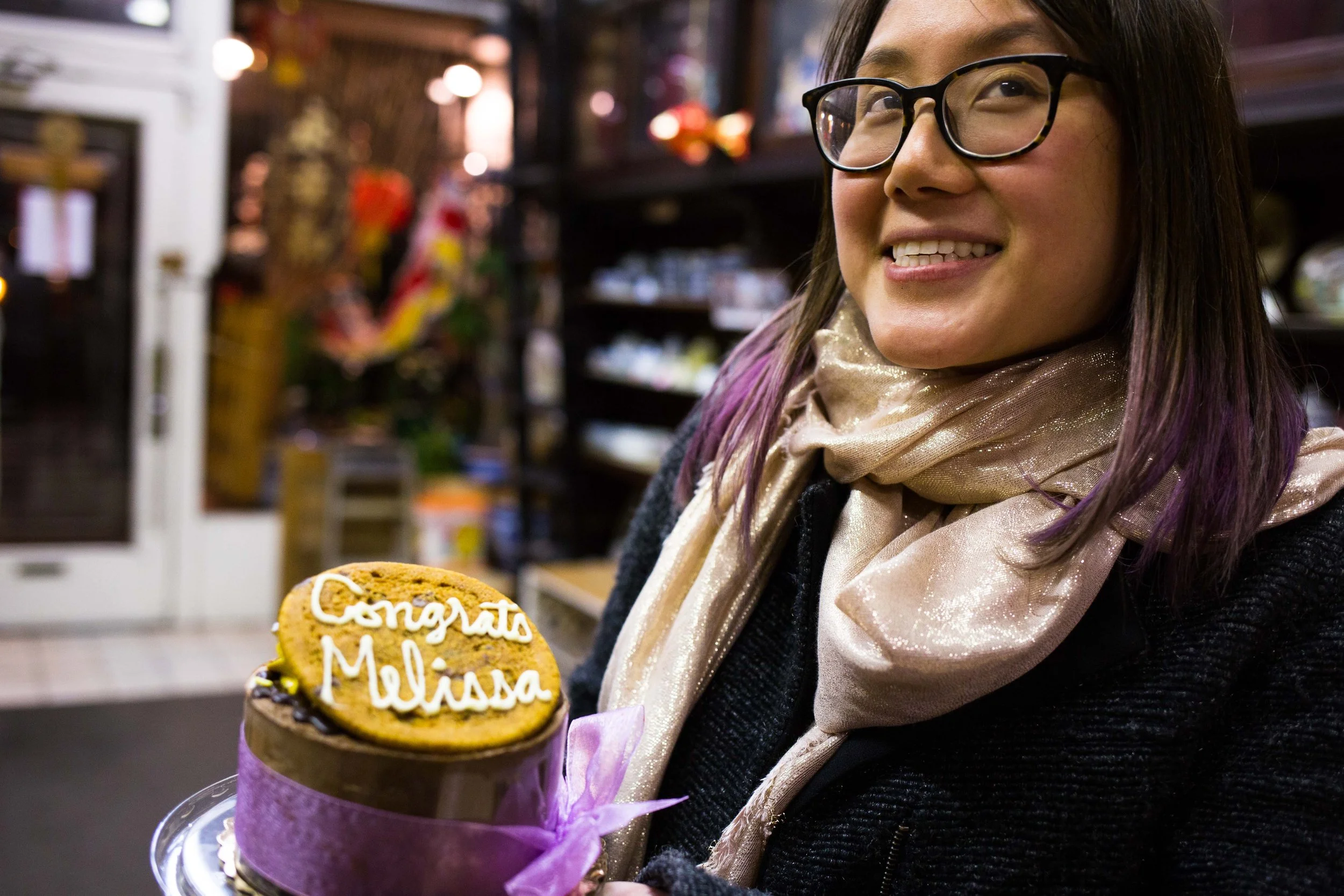 A woman with glasses and a beige scarf holding a birthday cake with a cookie topping that reads 'Congrats Melissa' in icing, inside a store.