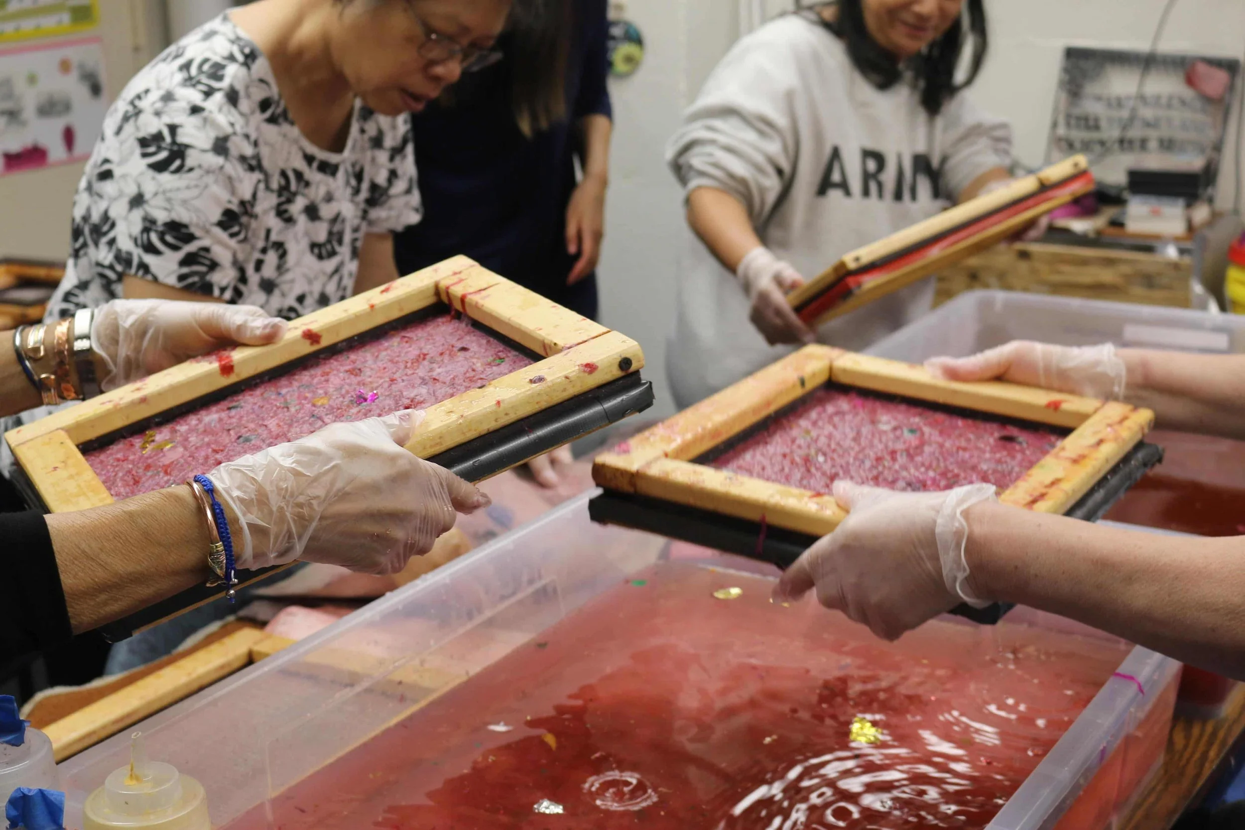 People working with screen printing frames to create screen-printed designs over a red ink on a table.