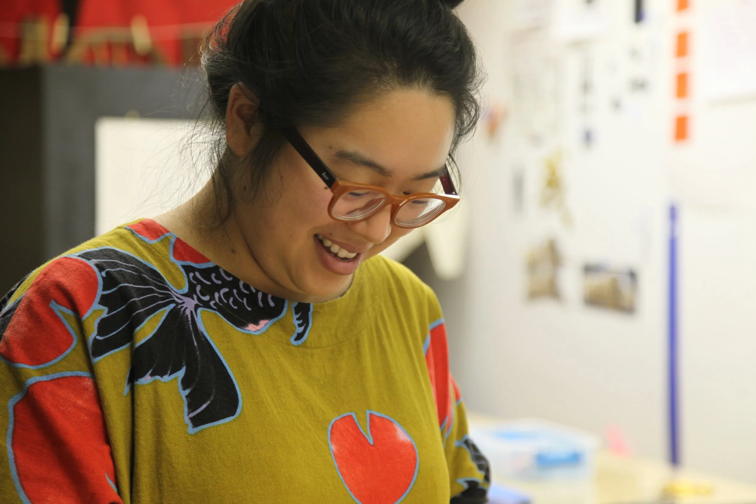 A woman with glasses smiling while looking down, wearing a colorful shirt with a fish design, in an indoor setting.