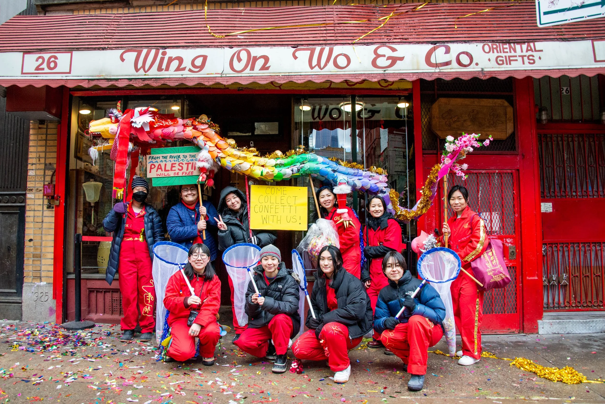 Group of ten women dressed in red uniforms and black jackets, holding butterfly nets, standing in front of a store with a colorful homemade dragon float and signs, celebrating a festival with confetti on the ground.