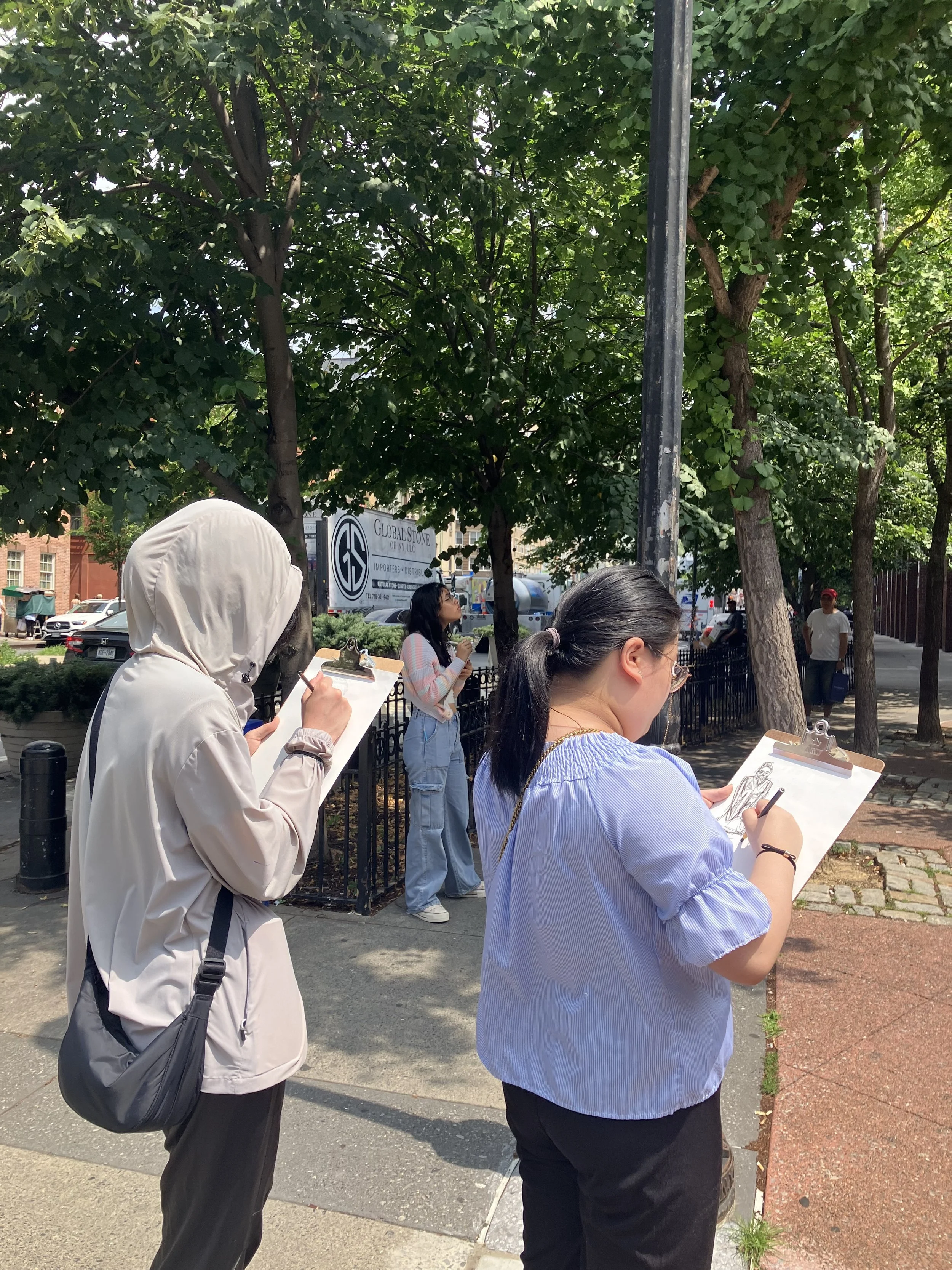 Three women drawing on clipboards while standing outdoors on a sidewalk under trees, with a street and parking lot in the background.