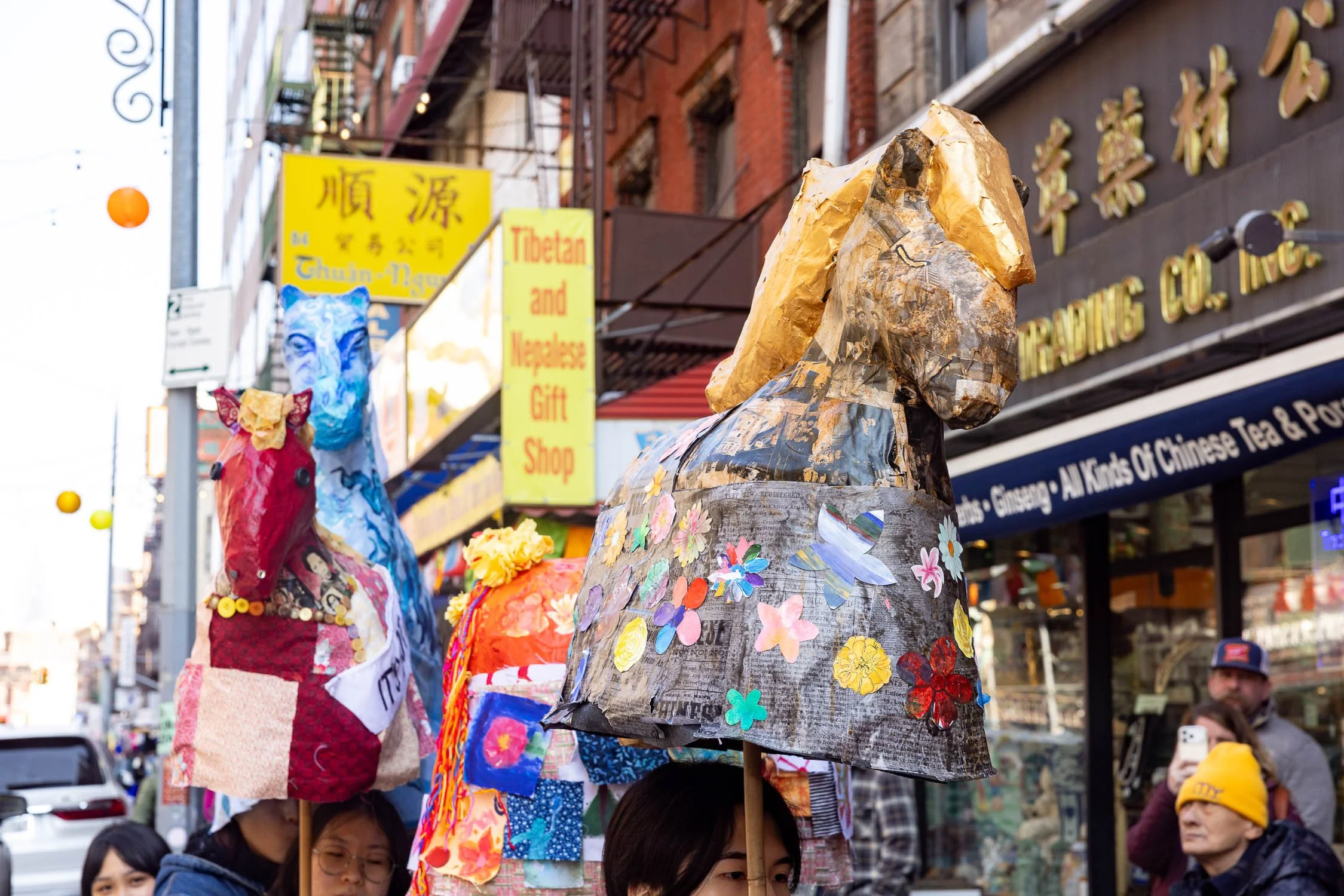 Fire horse puppet, Michelle parading through Chinatown. Michelle has a golden coat and mane. Her body is made of a collage of Chinese immigrant communities. Michelle's coat is made of sinophobic rhetoric and laws, but flowers emerge on top