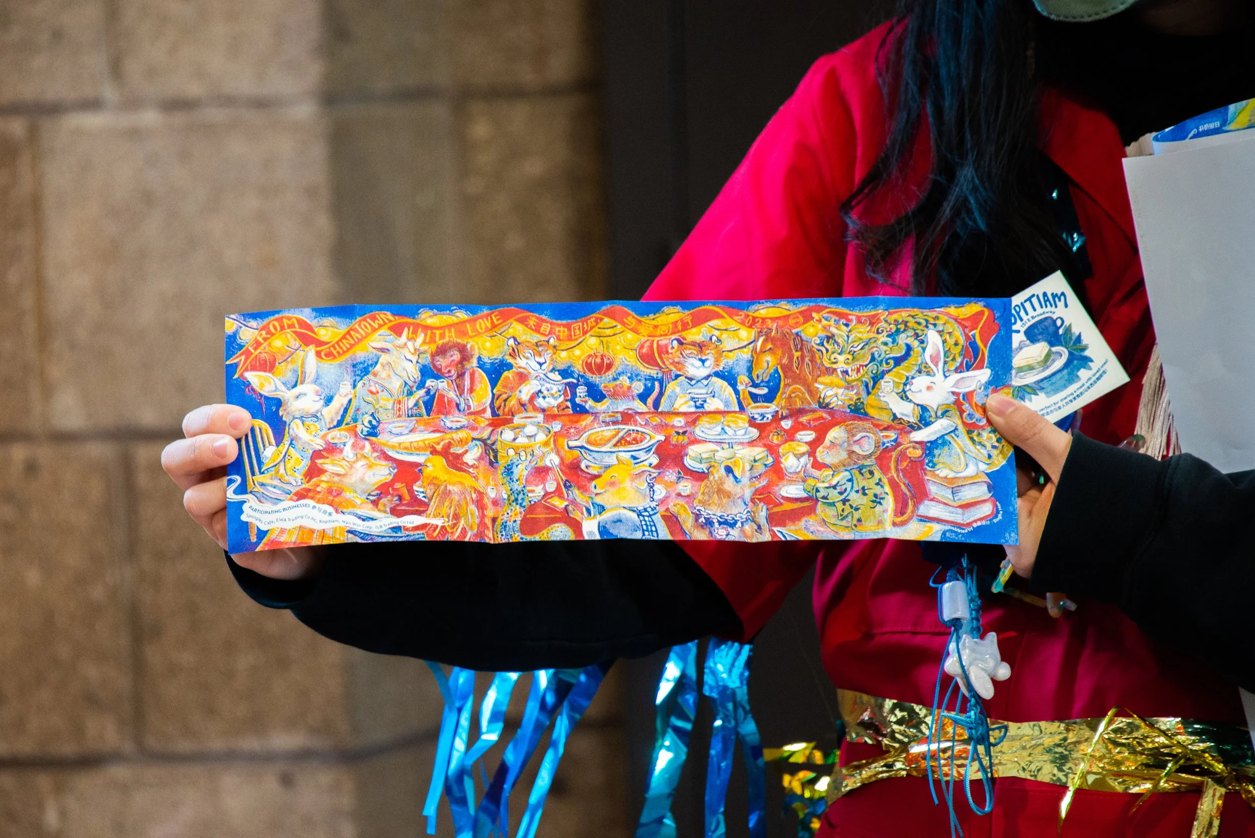 Person holding a colorful illustration of the animals from the Chinese Zodiac celebrating around a dinner table, with text that reads "From Chinatown with Love".