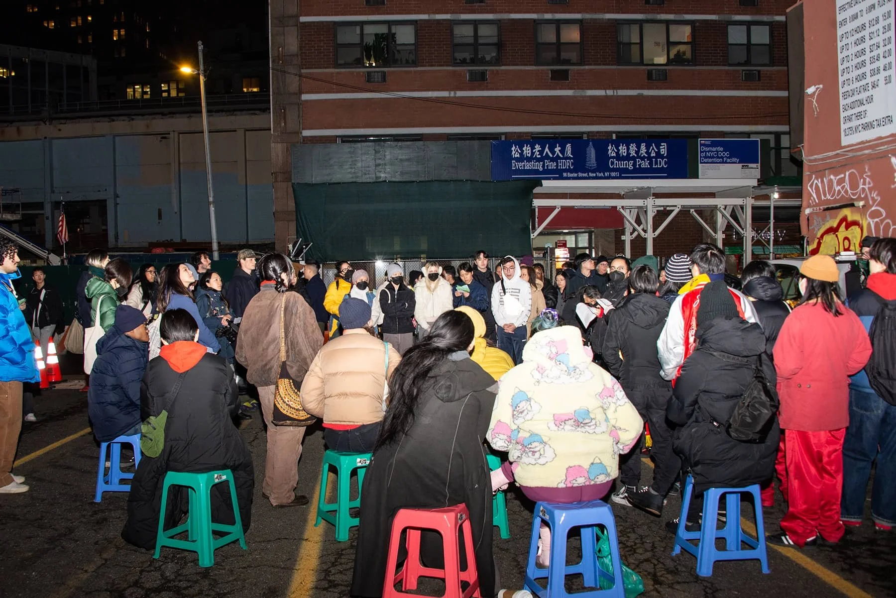 A large group of people gathered outdoors at night, with many seated on colorful stools and some standing, facing a speaker or presentation in front of a building with Chinese and English signs.